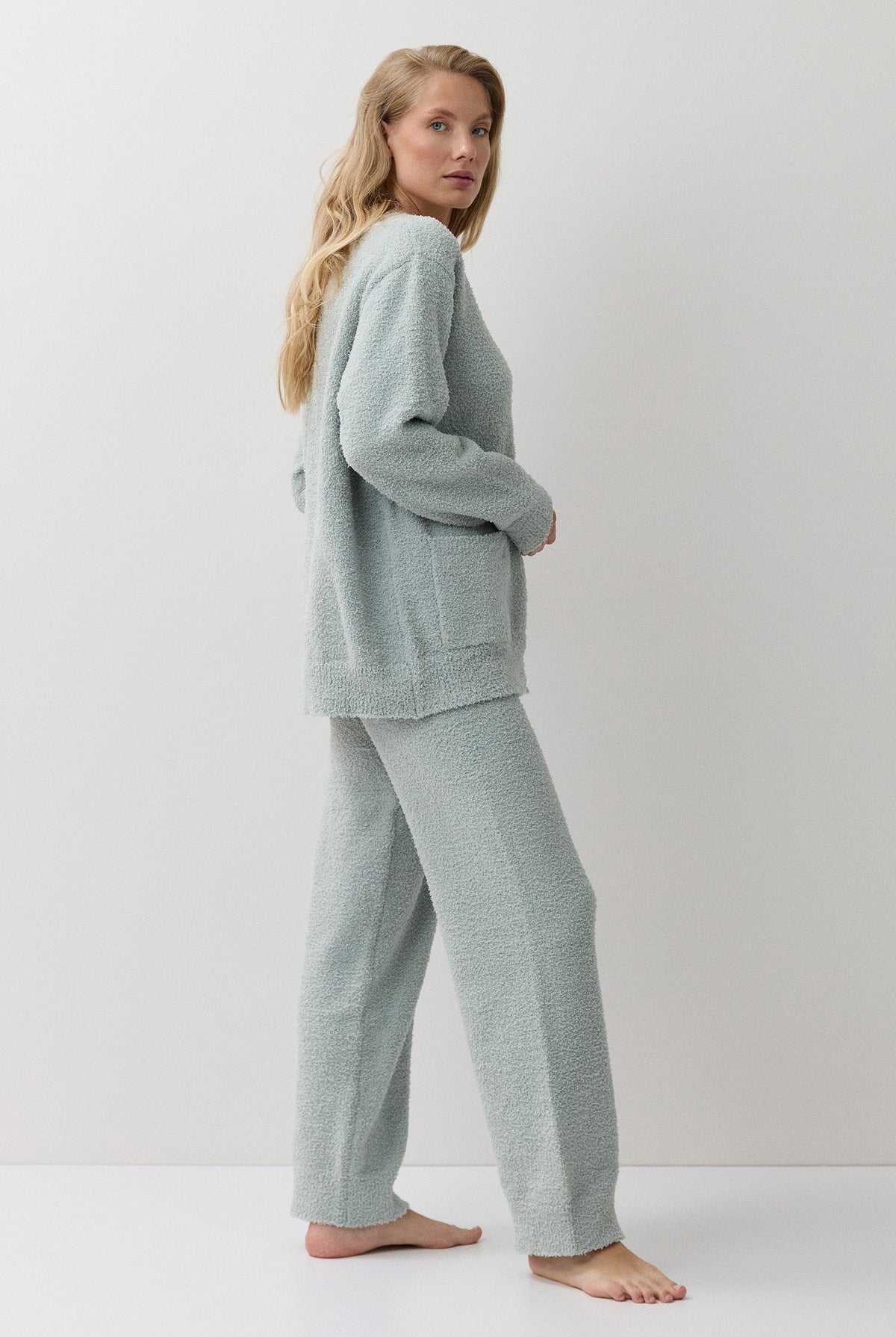 A woman stands barefoot in a studio wearing a matching light sage green loungewear set consisting of a long-sleeve knit top and wide-leg pants.