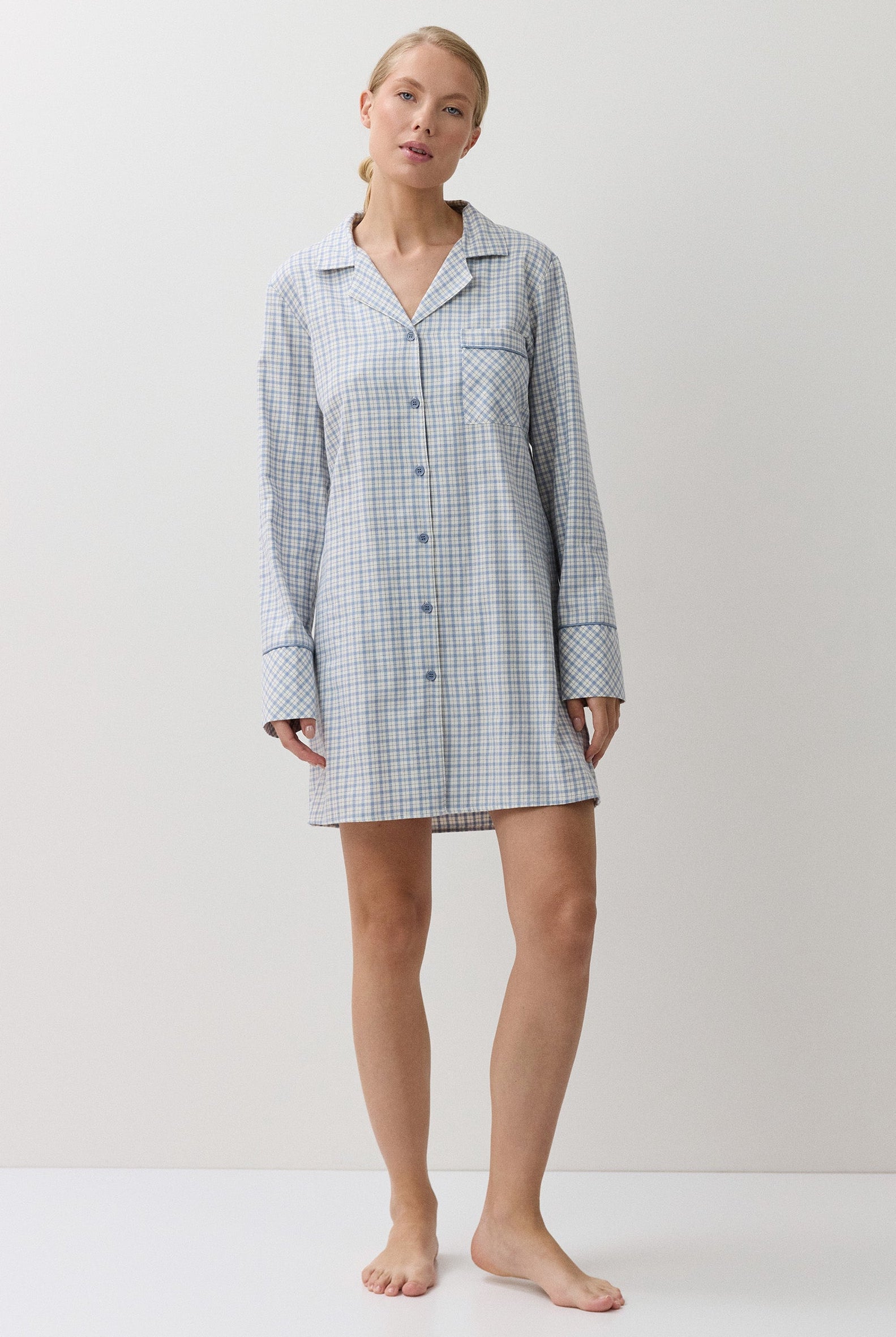 A woman modeling a light blue and white gingham check long-sleeve pajama shirt dress against a plain white studio background.