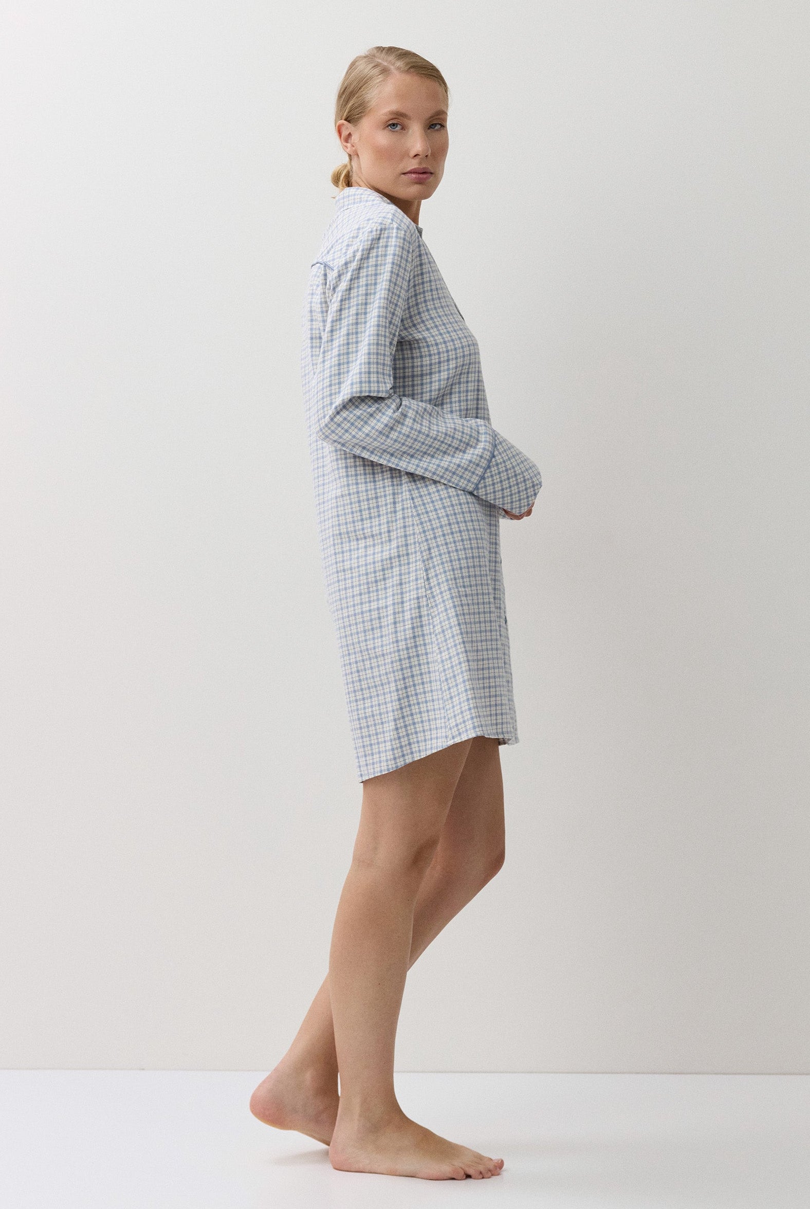 A woman stands barefoot in profile wearing a light blue and white gingham check long-sleeve robe against a plain off-white studio background.