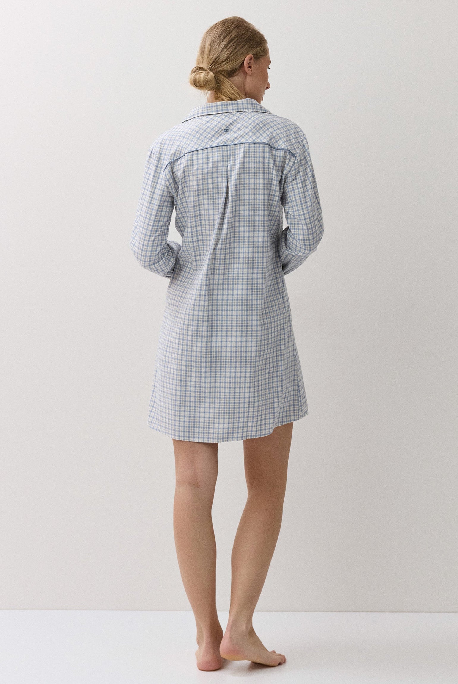 A woman stands barefoot from the back wearing a light blue and white gingham check long-sleeve shirt dress or pajama shirt against a plain off-white studio background.