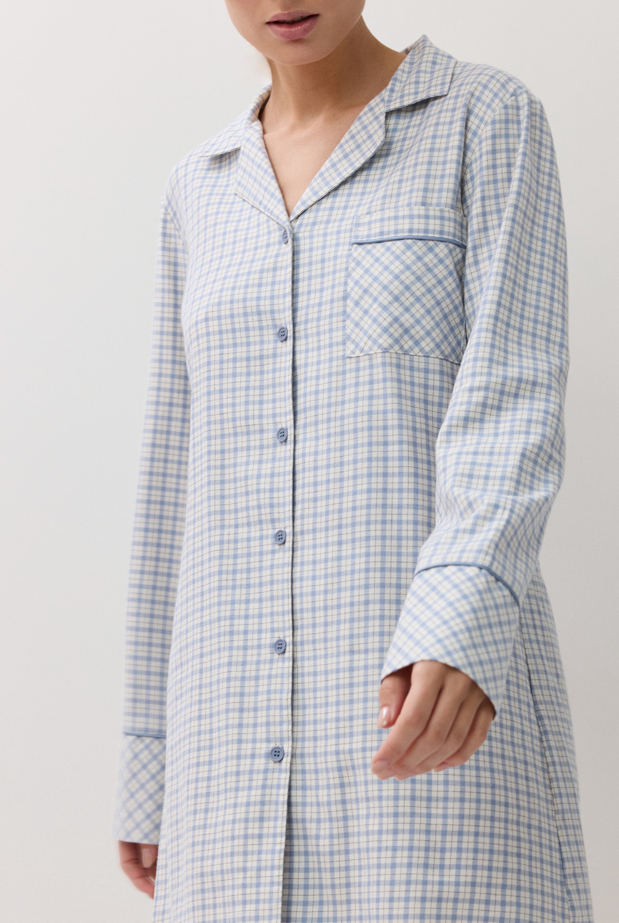 A woman wearing a light blue and white checkered long-sleeve button-up nightshirt, shown from the neck down against a plain light background.
