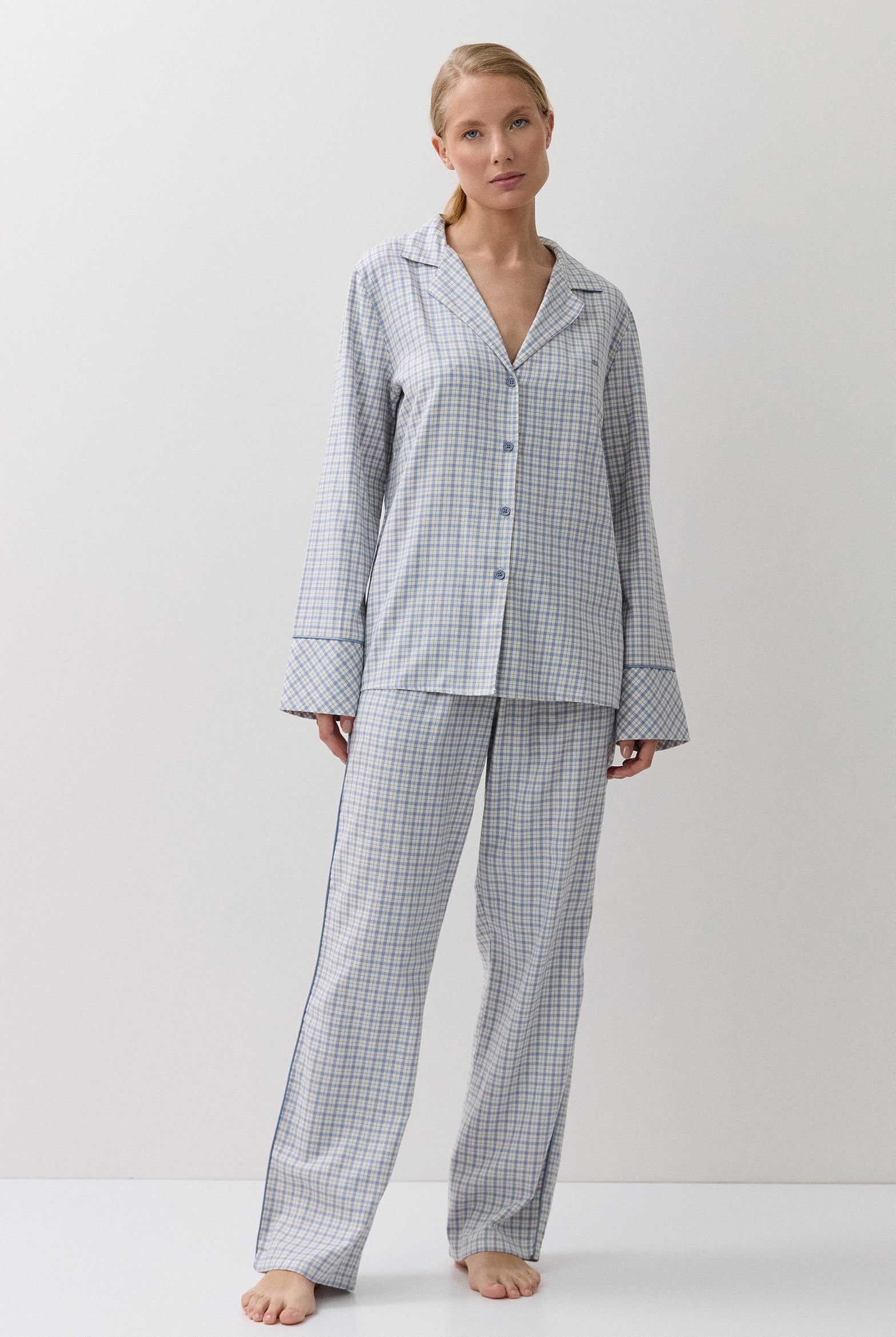 A woman stands barefoot in a studio wearing a matching long-sleeve button-front pajama top and wide-leg pajama pants in a small blue and white gingham check pattern.