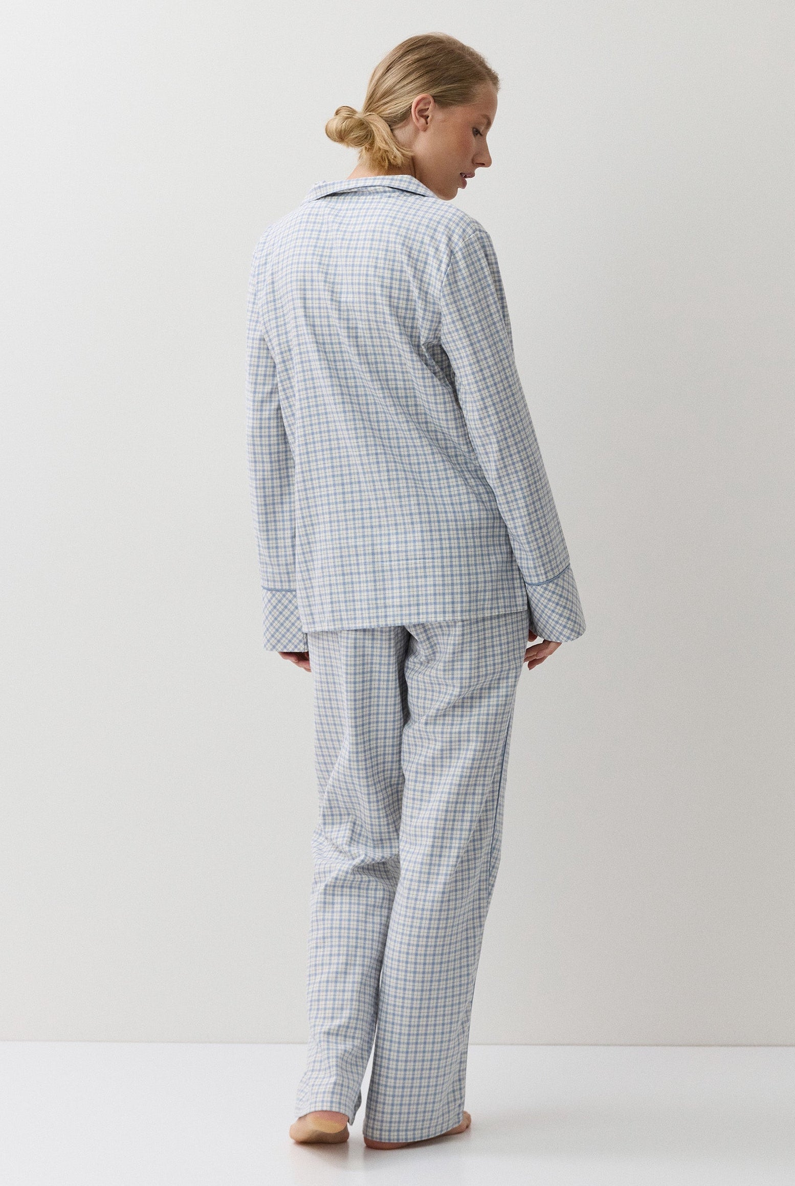 A woman stands with her back to the camera wearing a light blue and white gingham pajama set, consisting of a long-sleeve button-up top and matching pants, in a minimalist studio setting.