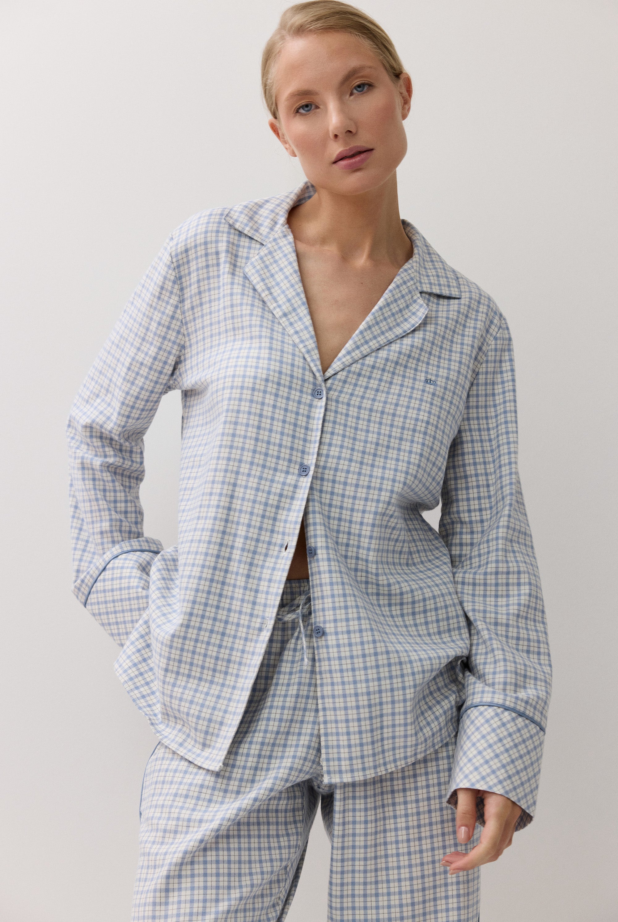A woman modeling a light blue and white checkered pajama set, featuring a long-sleeve button-up shirt and matching pants, against a plain white background.