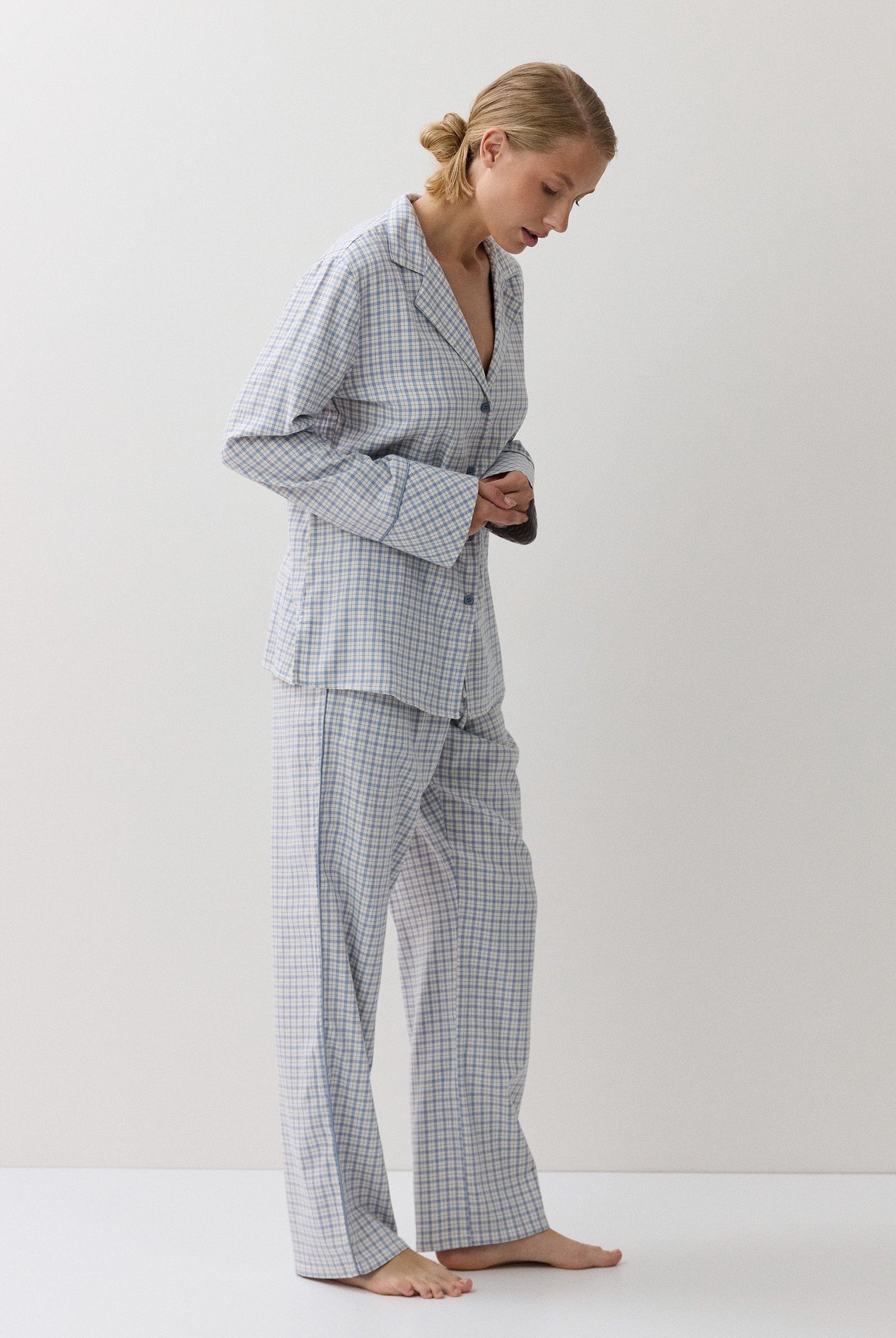A woman wearing a light blue and white gingham pajama set, standing barefoot against a plain white studio background.