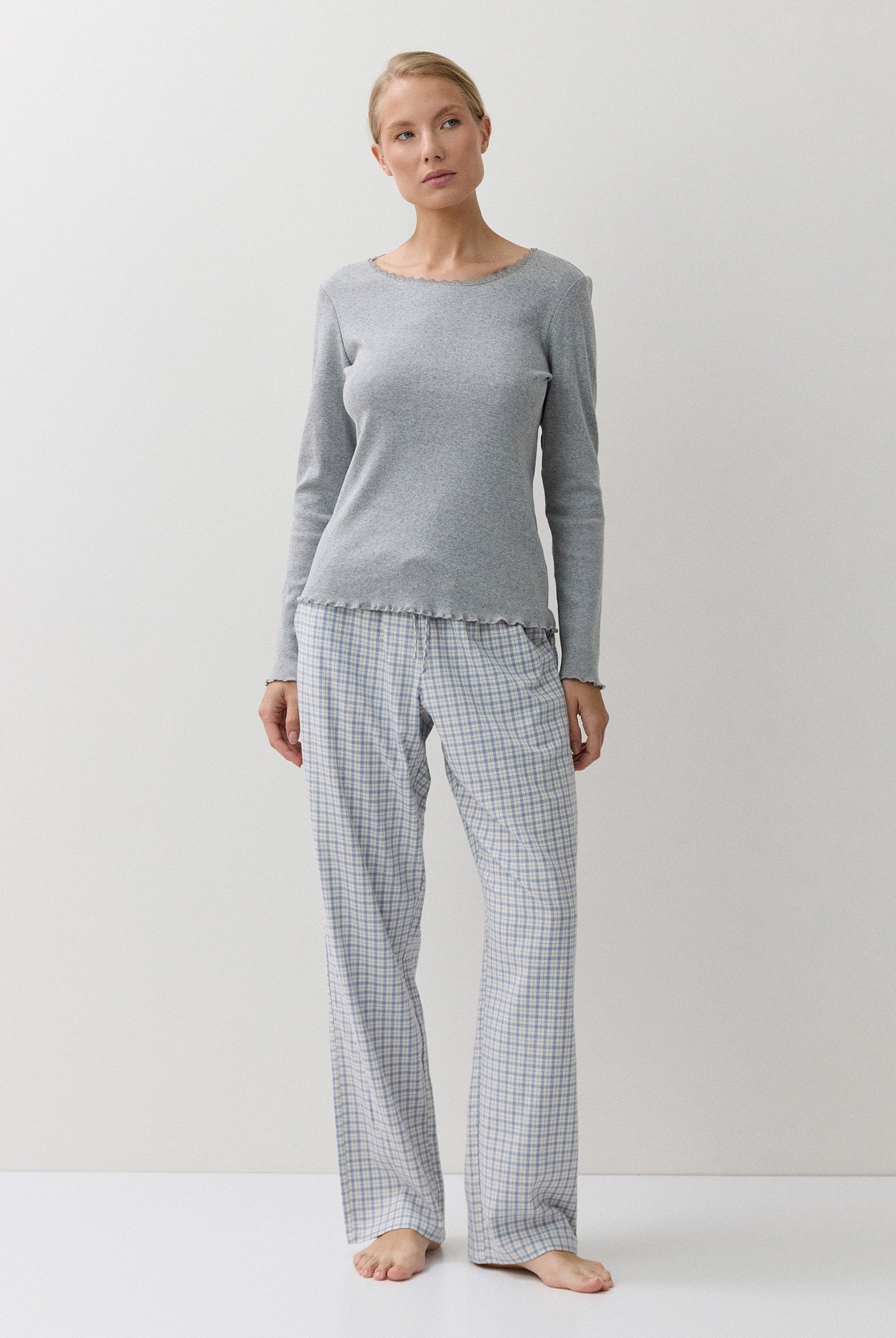 A woman stands barefoot in a studio wearing a light gray long-sleeve top and matching gray-and-white gingham pajama pants against a plain light background.