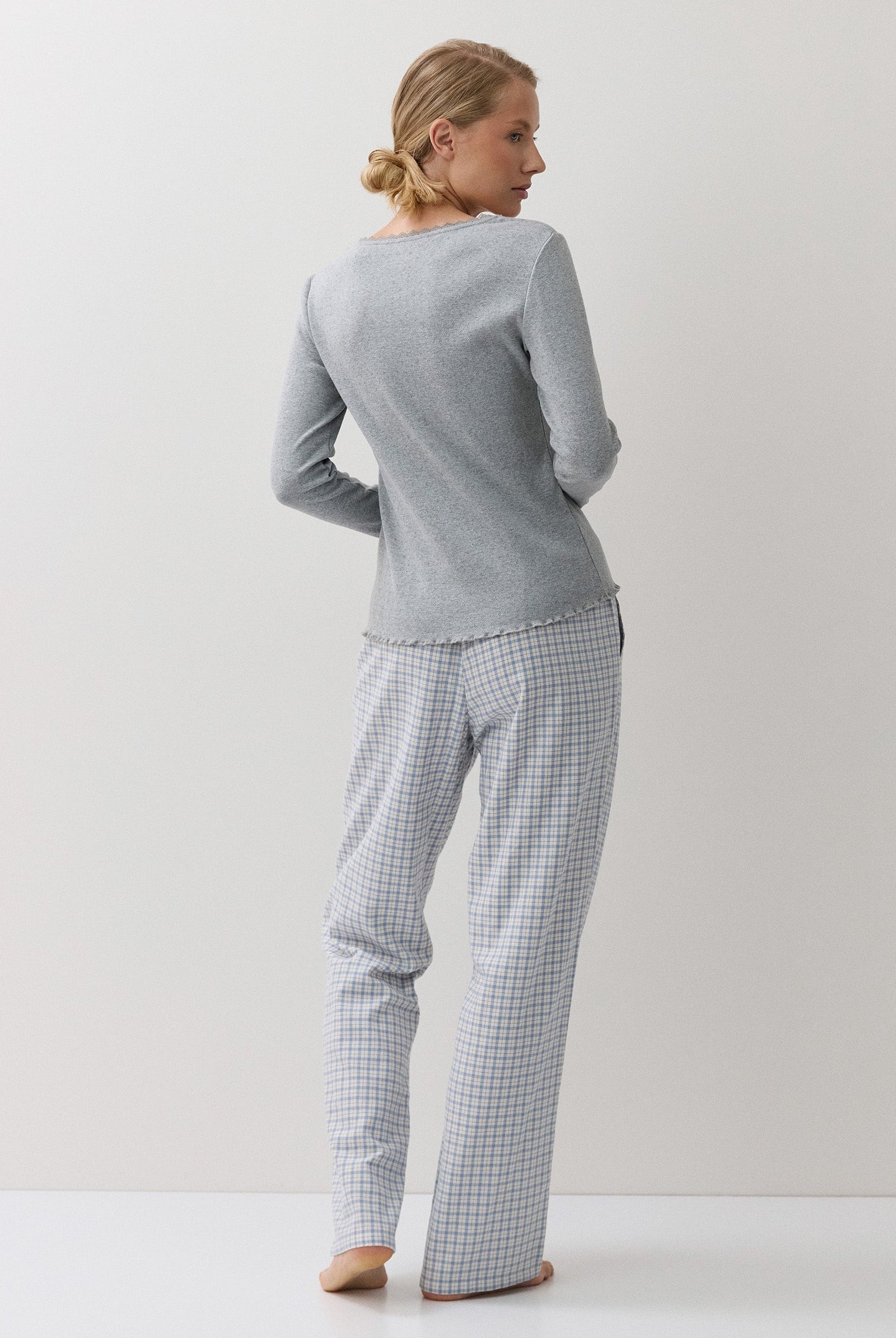A woman stands barefoot against a plain white studio background, viewed from the back, wearing a light gray long-sleeve top and matching white-and-blue checkered pajama pants.