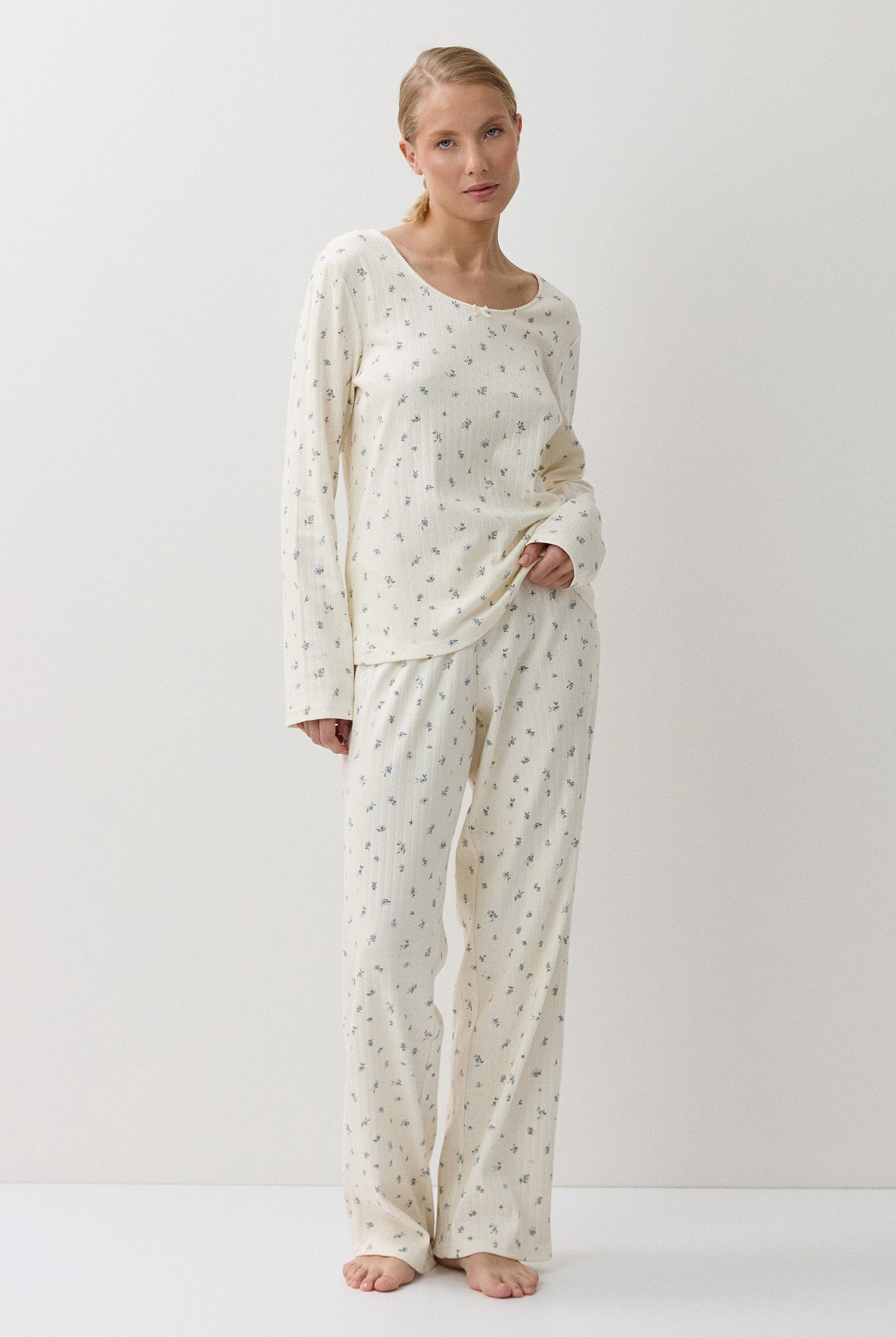 A woman modeling a two-piece long-sleeve pajama set with a small repeating floral pattern on an off-white base, standing against a plain white studio background.