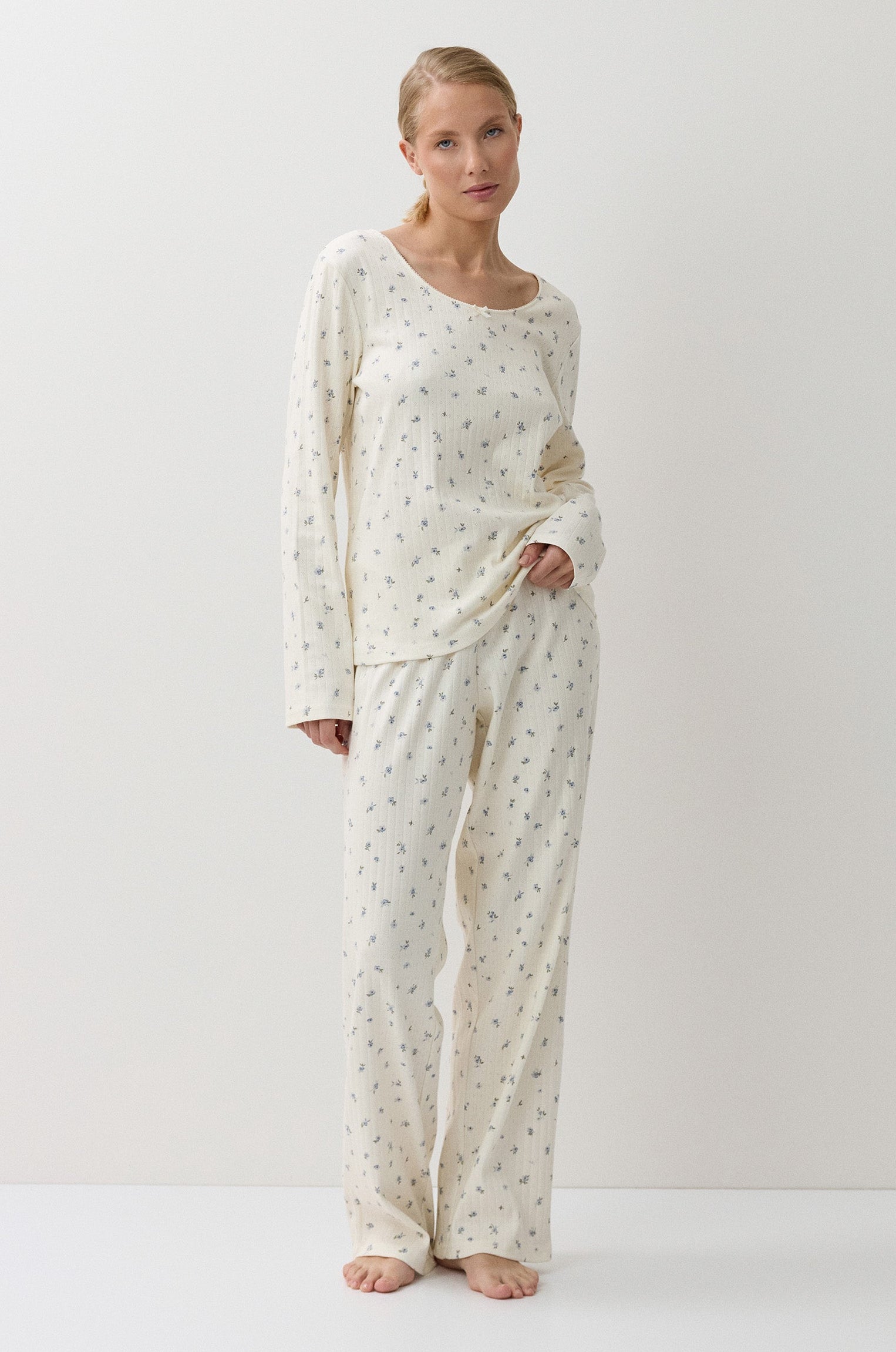 A woman modeling a two-piece long-sleeve pajama set with a small repeating floral pattern on an off-white base, standing against a plain white studio background.