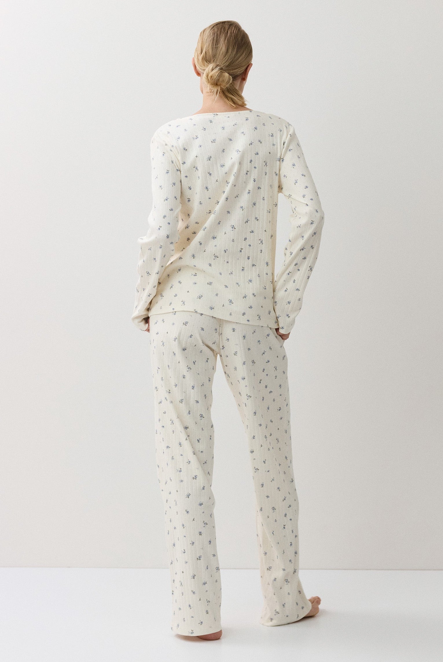 A woman stands with her back to the camera wearing a matching long-sleeve pajama top and pants set in a warm off-white fabric with a small, repeating floral print.