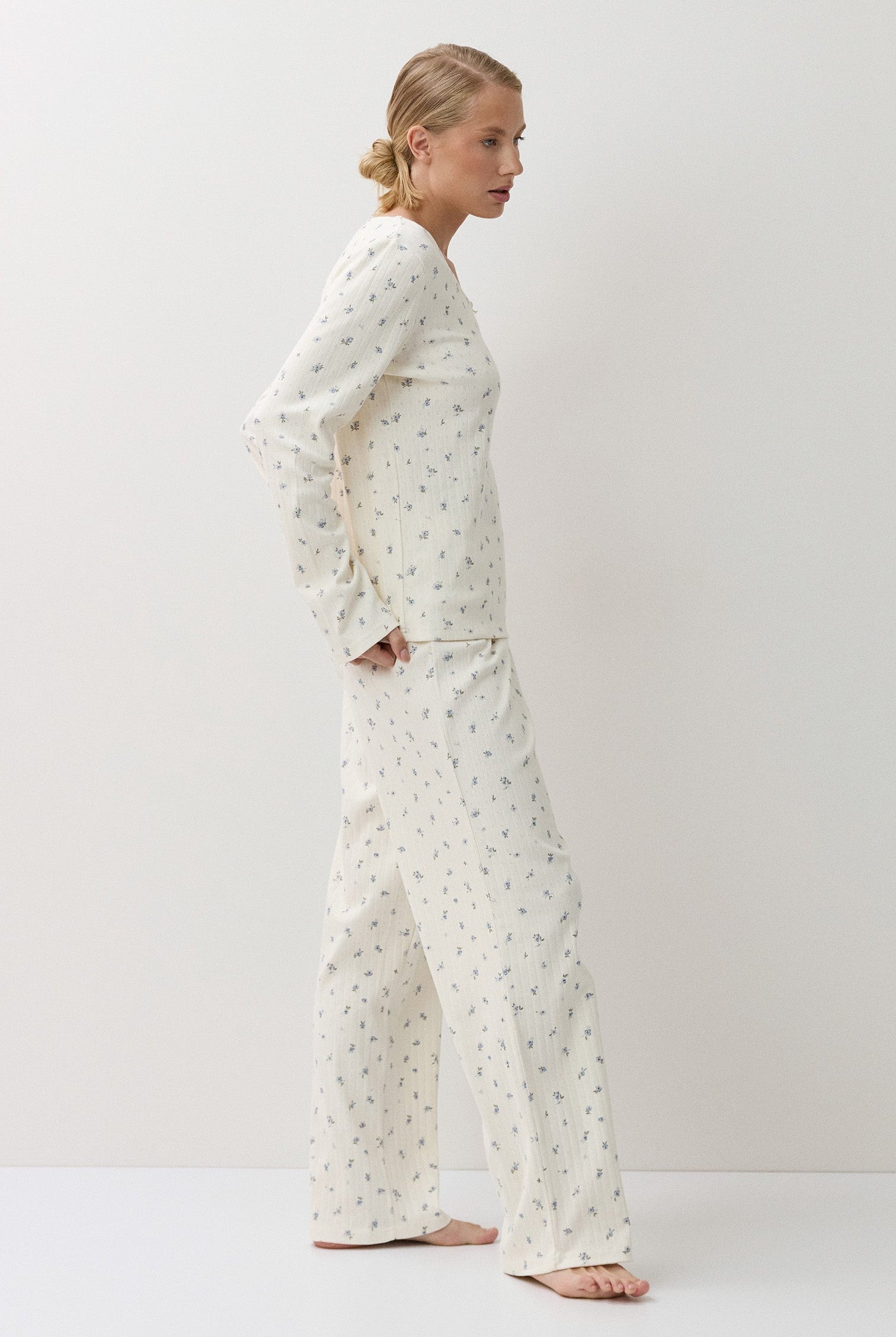 A woman stands in profile wearing a matching long-sleeve pajama top and wide-leg pants set in a warm off-white fabric with a small, repeating green floral print. The photo is taken in a minimalist studio setting with a light white background.