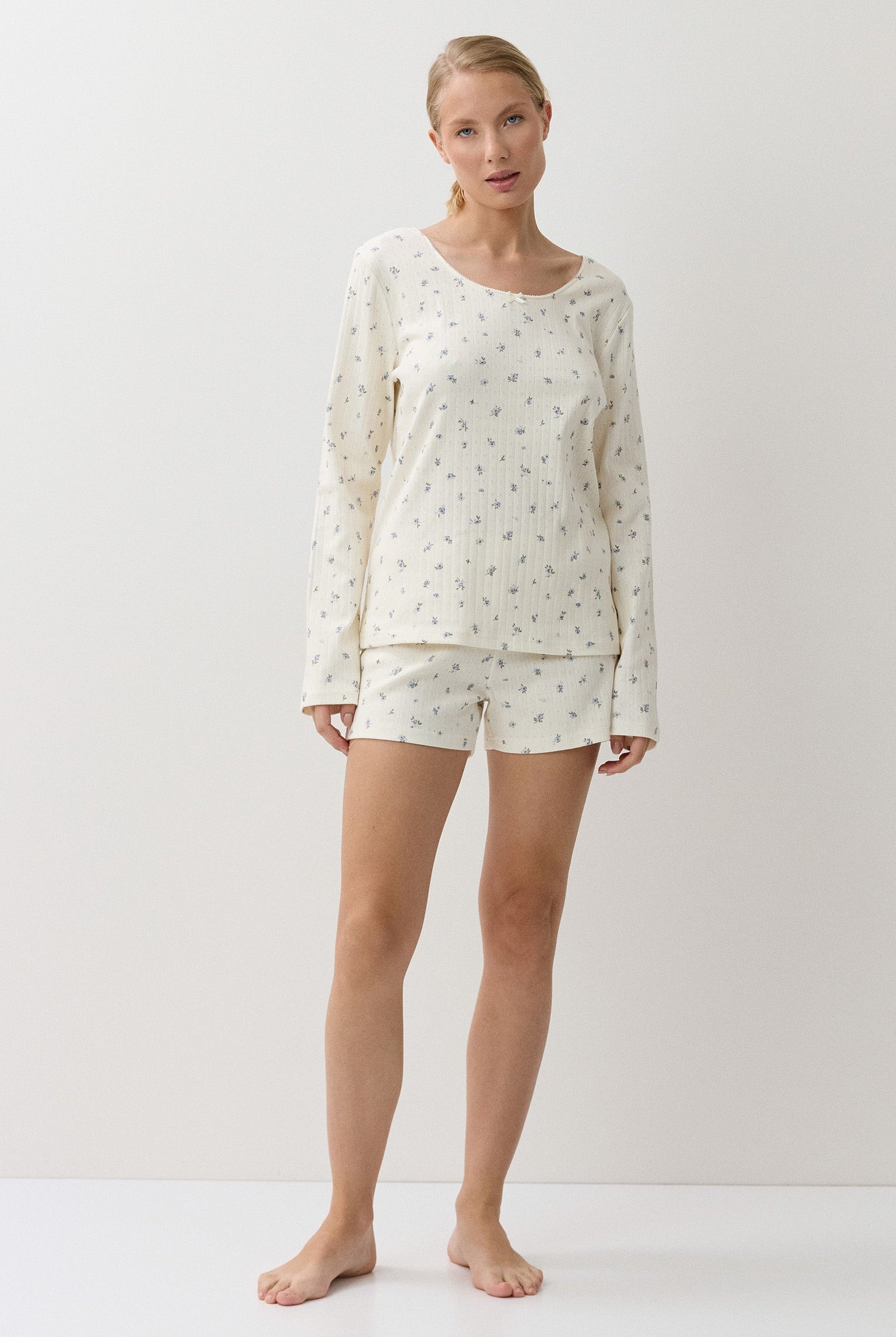 A woman modeling a two-piece pajama set consisting of a long-sleeve top and matching shorts, both in an off-white fabric with a small floral pattern, set against a plain white studio background.