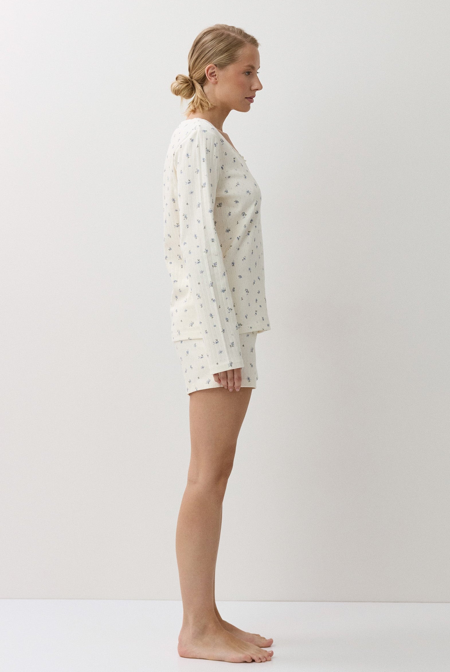 A woman stands in profile wearing a matching long-sleeve pajama top and shorts set in a warm off-white fabric with a small, scattered floral print. The image is shot in a minimalist studio setting with a warm, off-white background.
