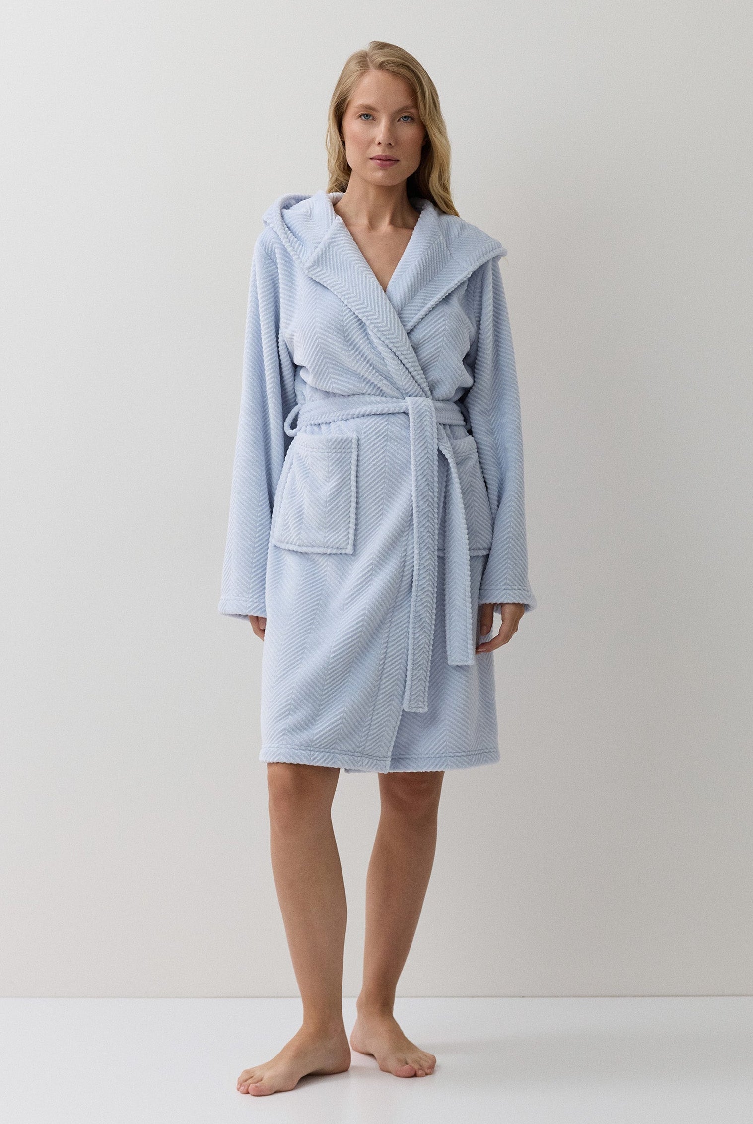 A woman modeling a light blue hooded bathrobe with a tie belt and front patch pocket, standing against a plain off-white studio background.