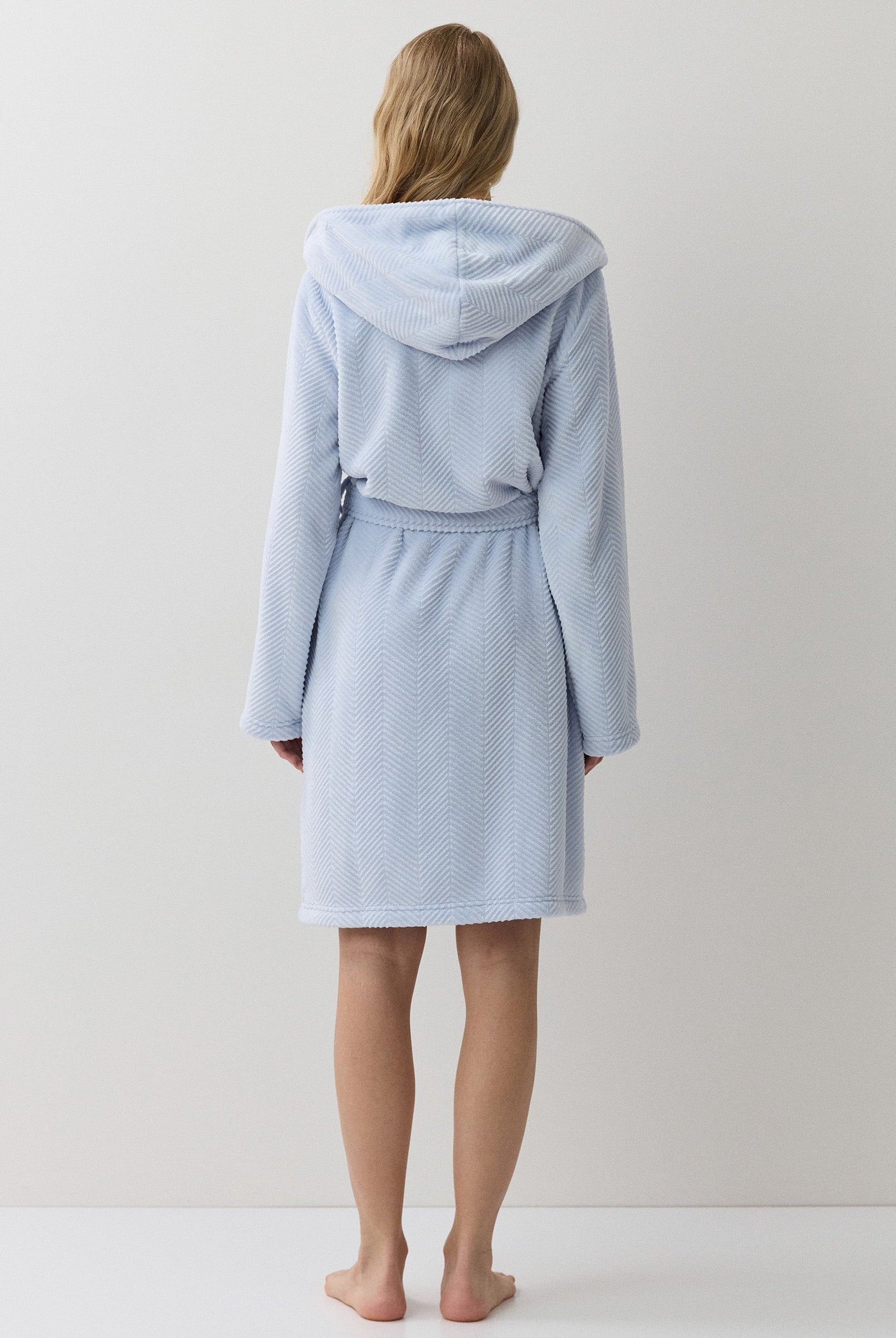 A woman stands with her back to the camera wearing a light blue hooded bathrobe against a plain white studio background.