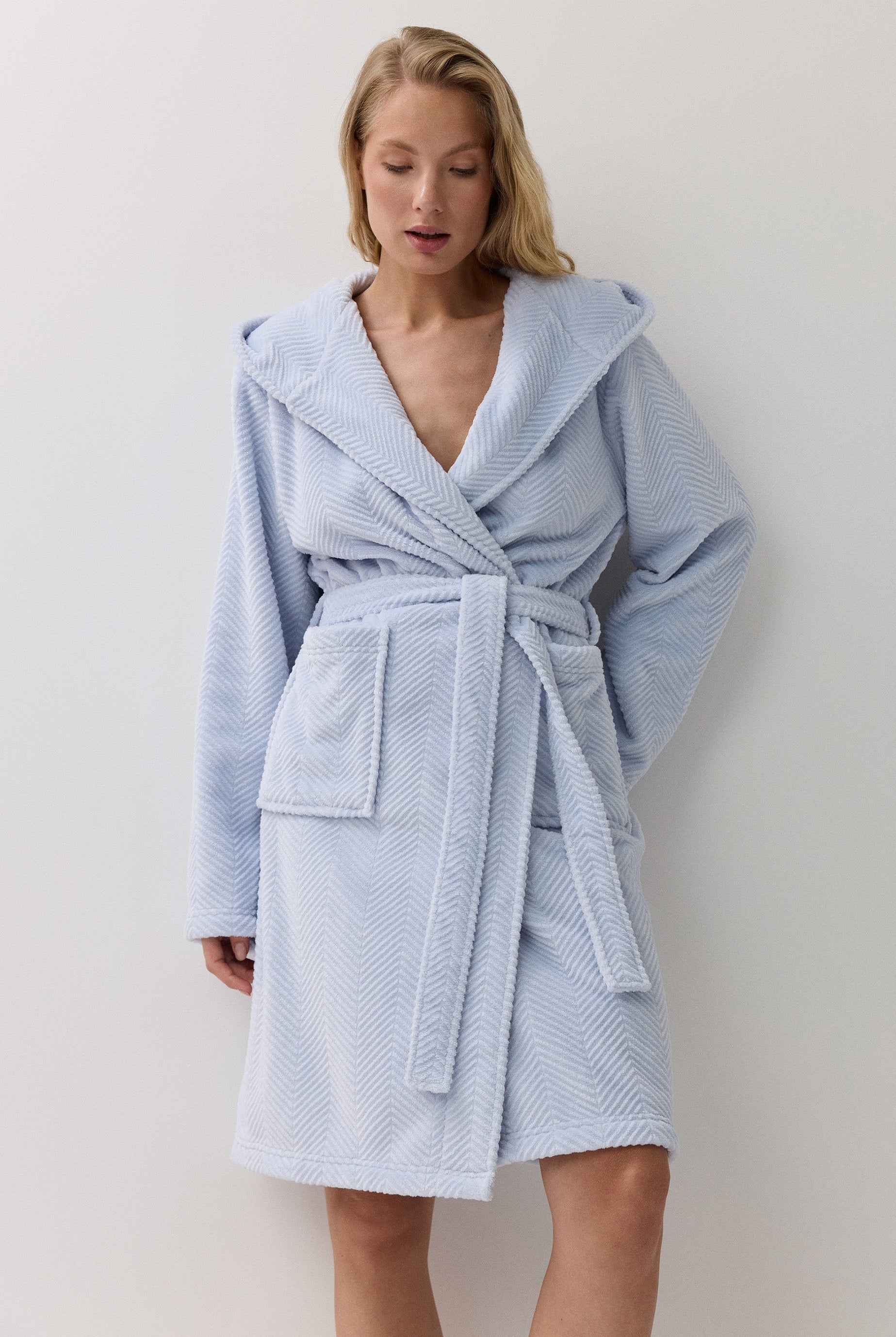 A woman wearing a light blue textured bathrobe with a hood, waist tie, and front patch pockets, standing against a plain white wall.