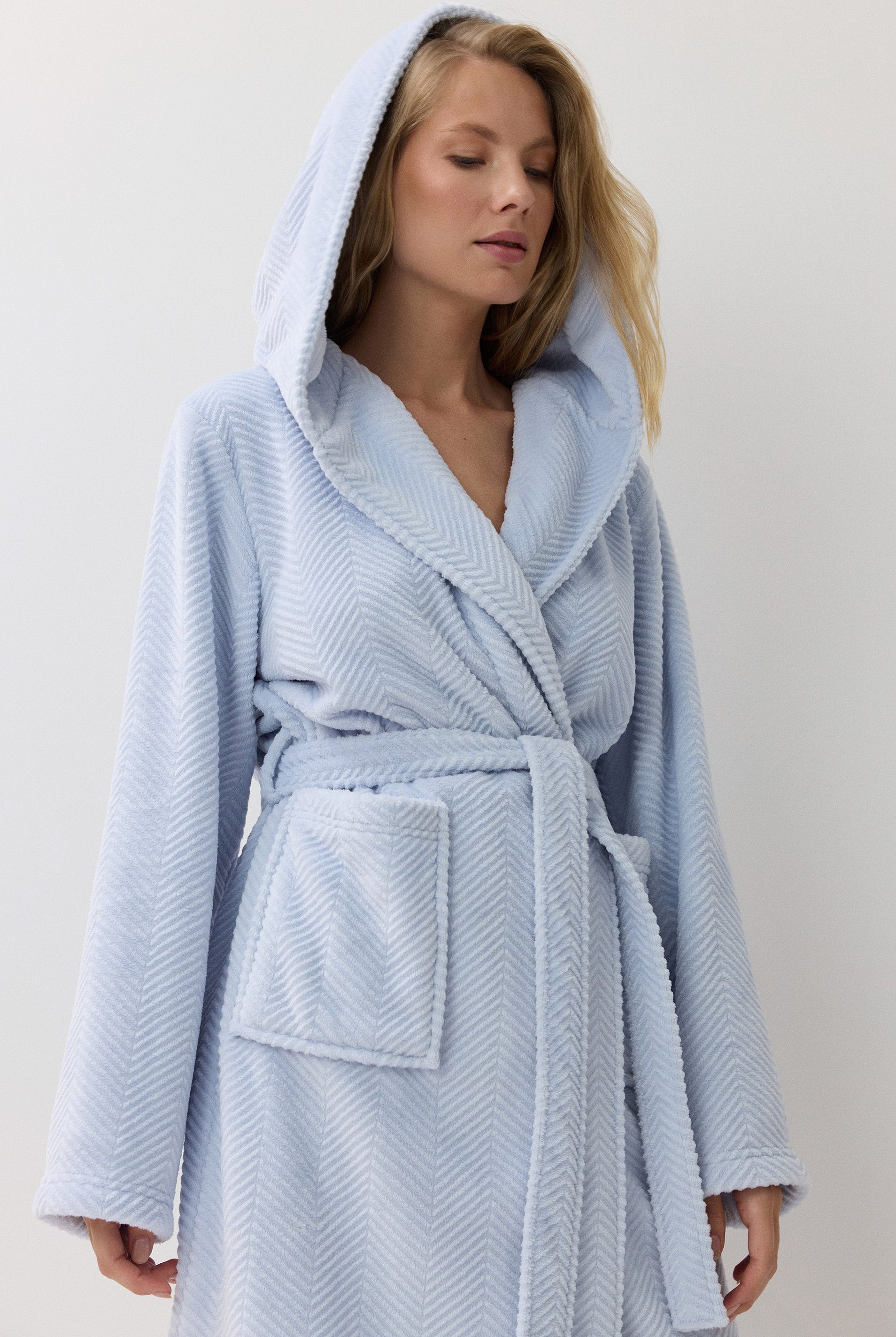 A woman wearing a light blue hooded bathrobe with a textured pattern, standing against a plain off-white background.