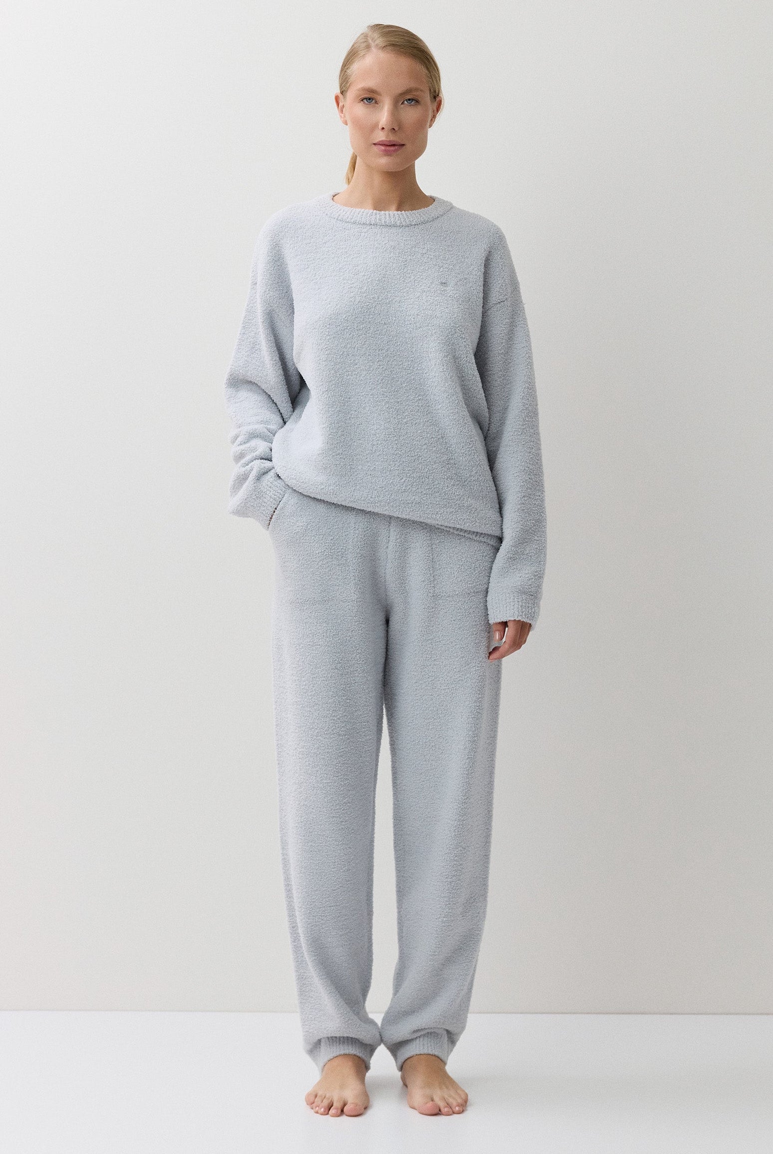A woman stands barefoot against a plain white studio background, wearing a light blue long-sleeve sweatshirt and matching jogger-style pants.