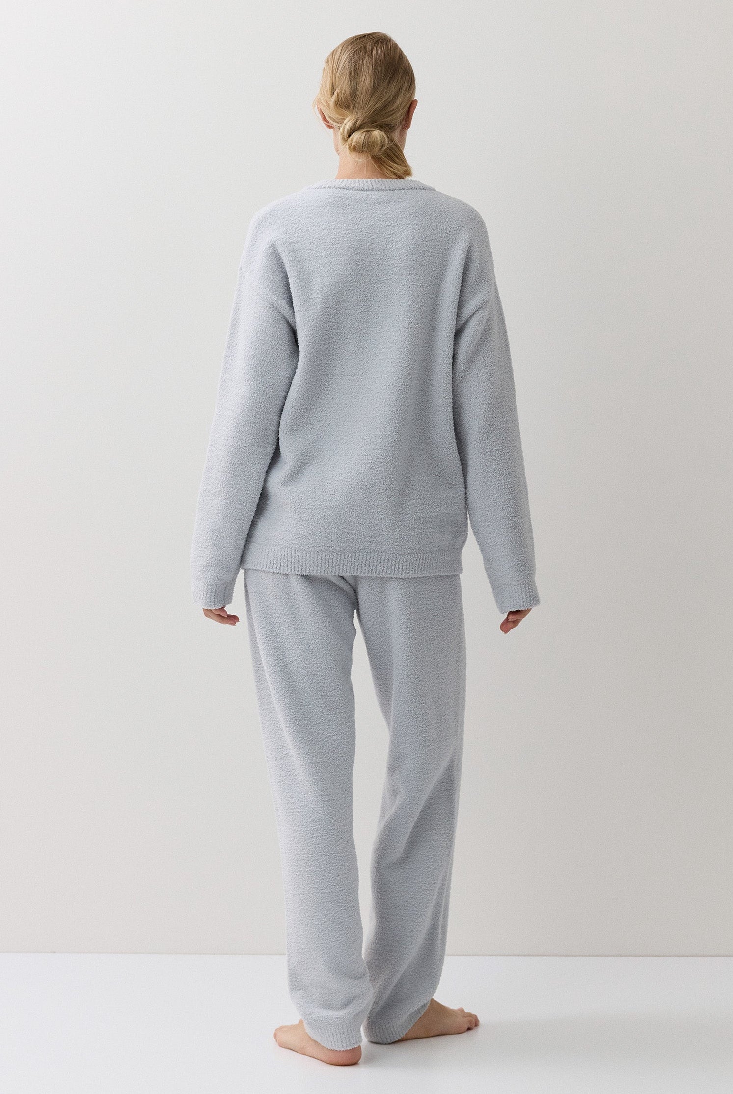 A woman stands with her back to the camera wearing a light blue lounge set consisting of a long-sleeve top and matching pants, set against a plain off-white studio background.