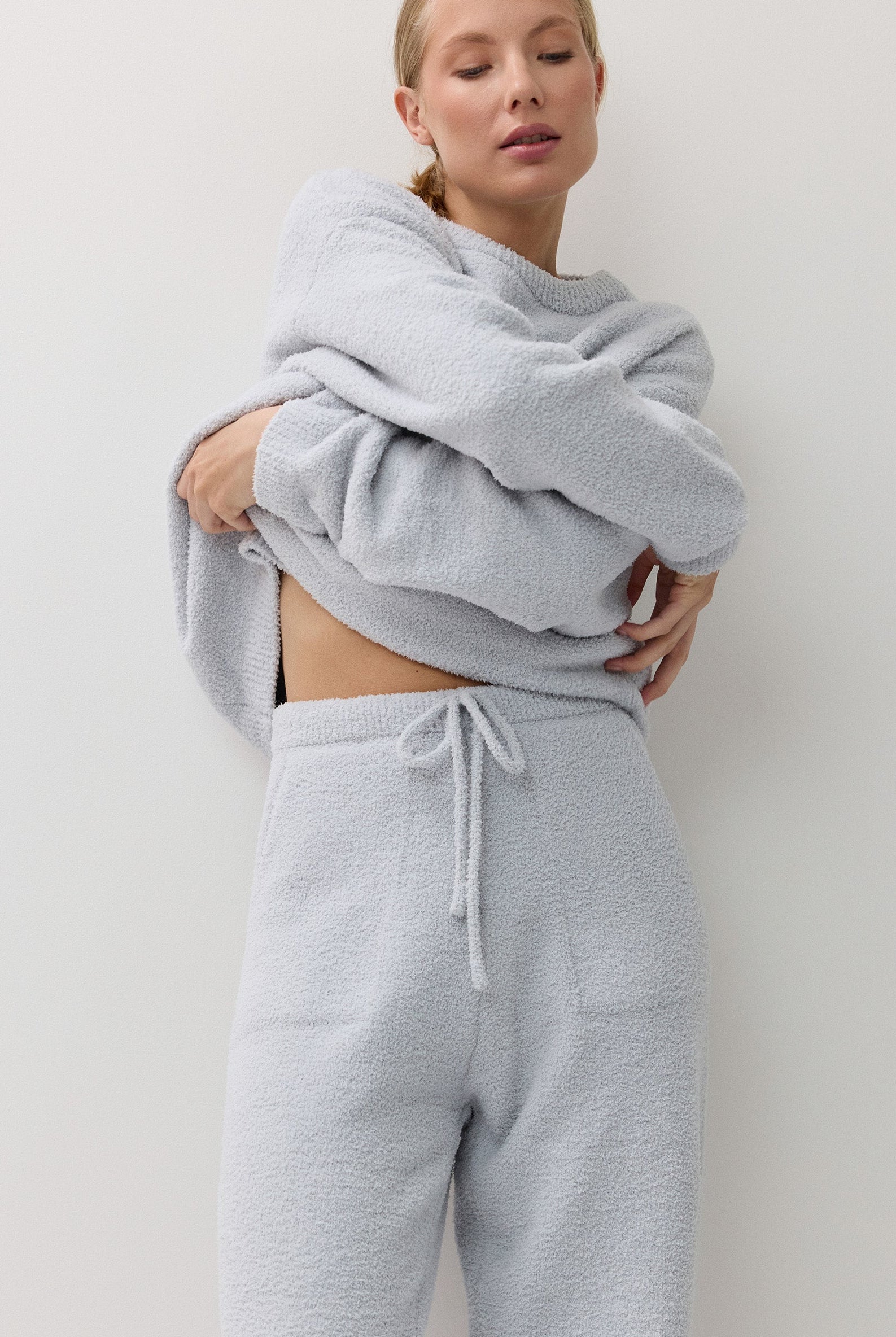 A woman modeling a light blue textured loungewear set consisting of a sweatshirt and matching drawstring sweatpants against a plain off-white background.