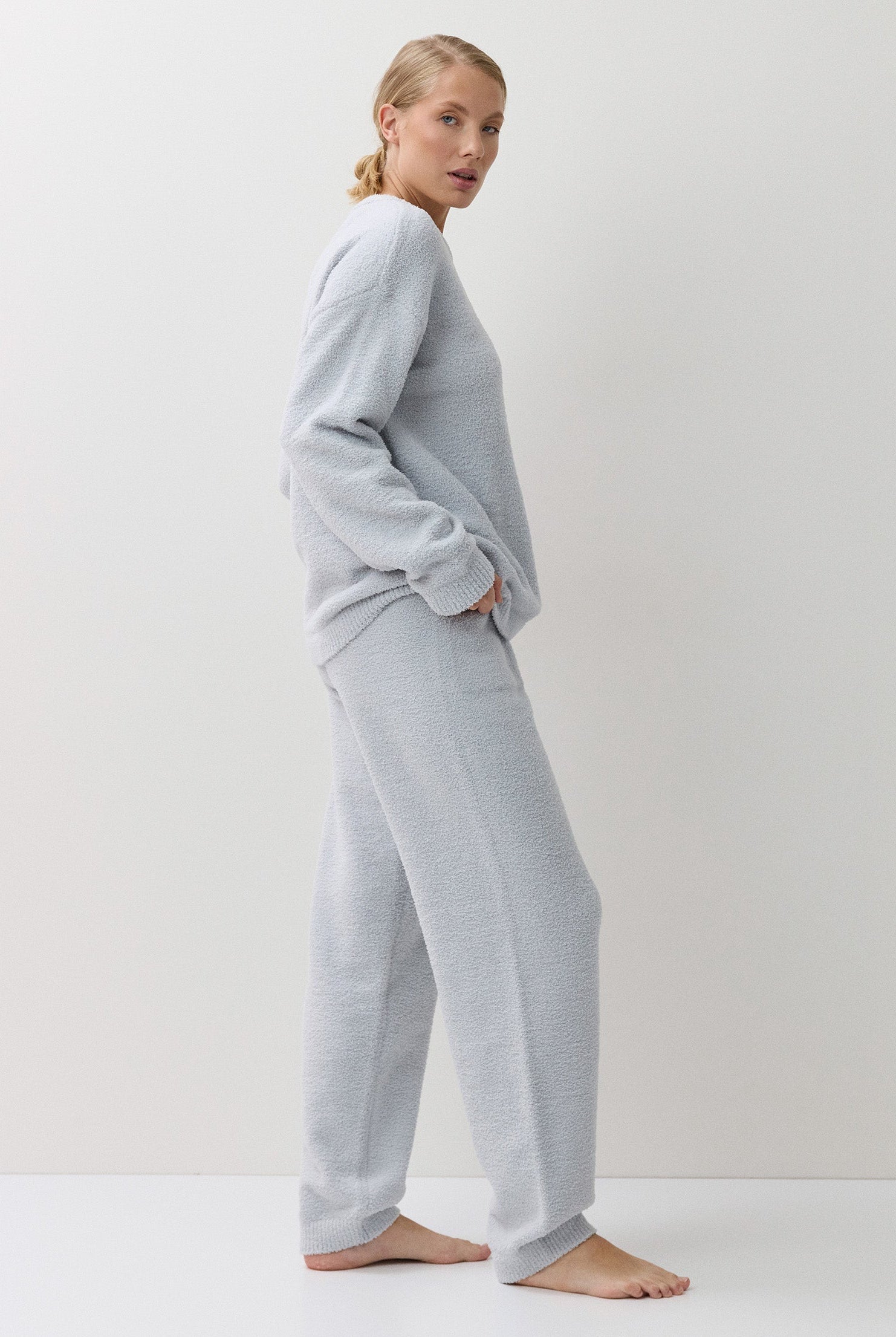 A woman stands barefoot in profile wearing a light blue two-piece lounge set consisting of a long-sleeve sweatshirt and matching wide-leg sweatpants against a plain white studio background.
