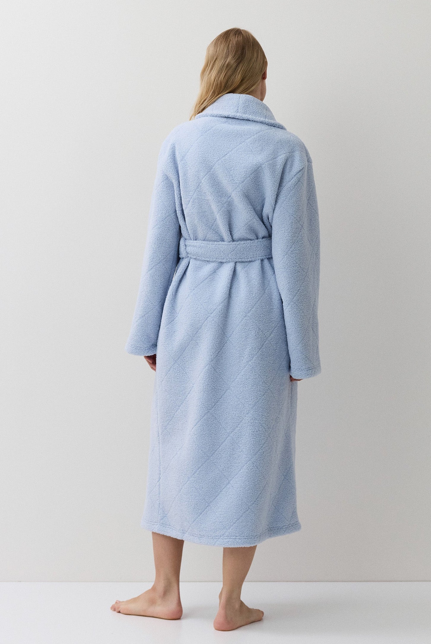 A woman stands with her back to the camera wearing a light blue bathrobe with a matching tie belt, set against a plain white studio background.