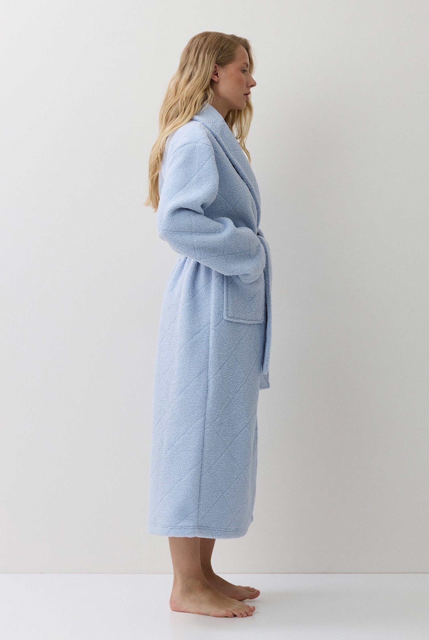 A woman stands in profile wearing a light blue bathrobe against a plain white studio background.