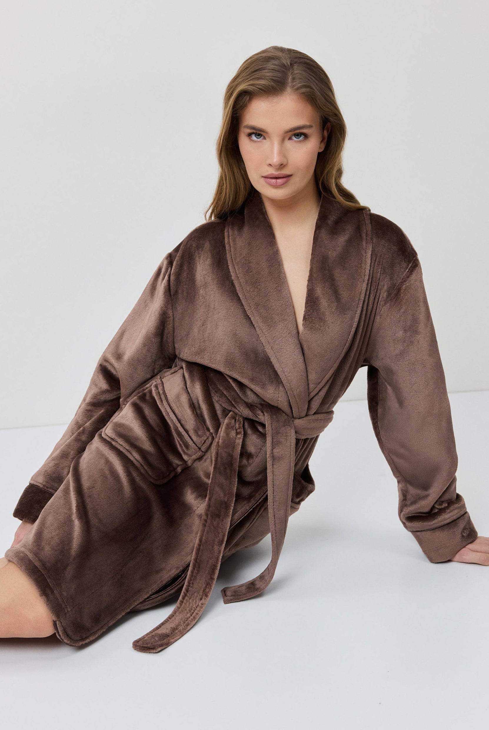 A woman wearing a plush, knee-length bathrobe from the back. The robe is a muted taupe-brown color with a soft, velvety texture and a matching waist tie.