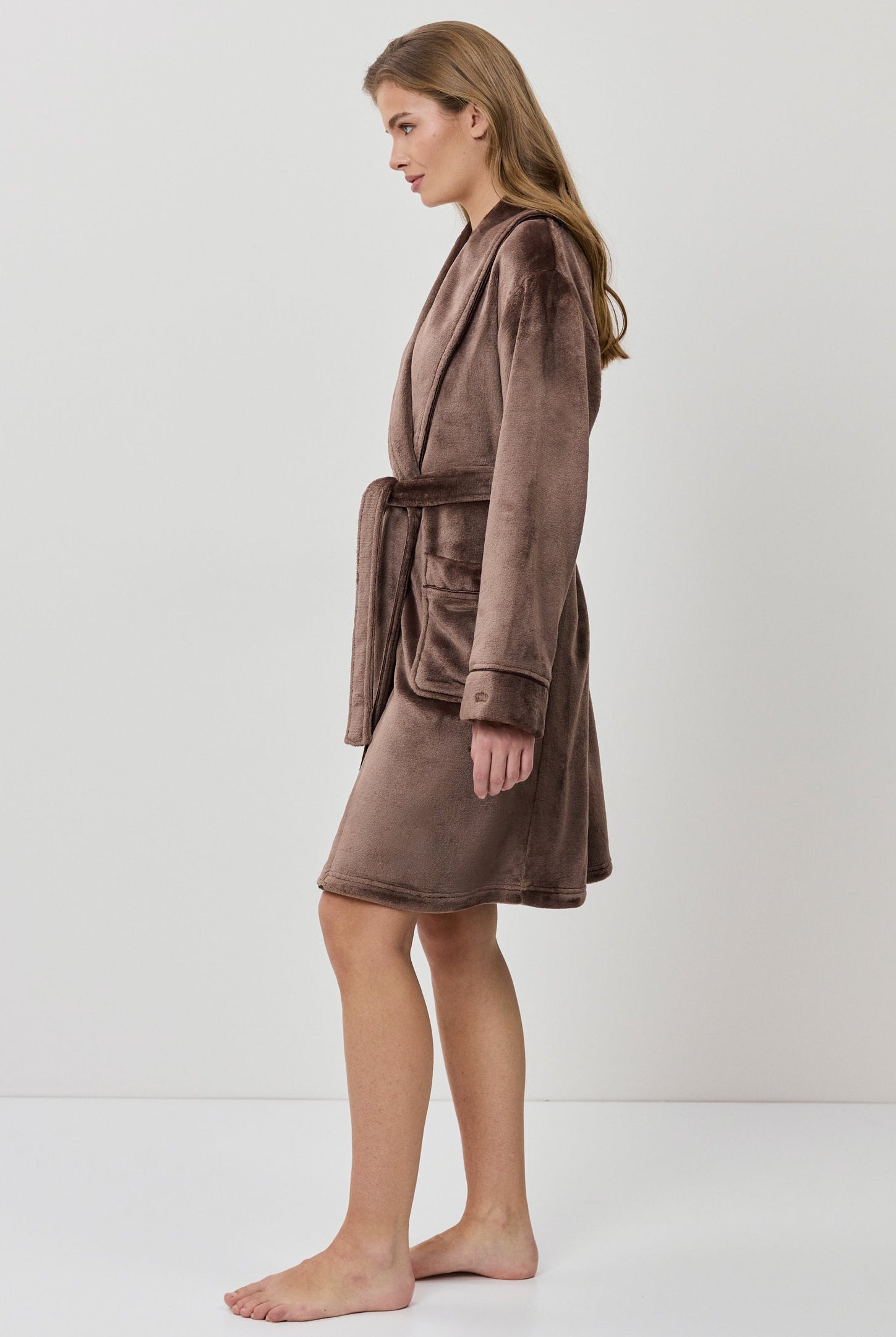 A woman wearing a plush, knee-length bathrobe from the back. The robe is a muted taupe-brown color with a soft, velvety texture and a matching waist tie.