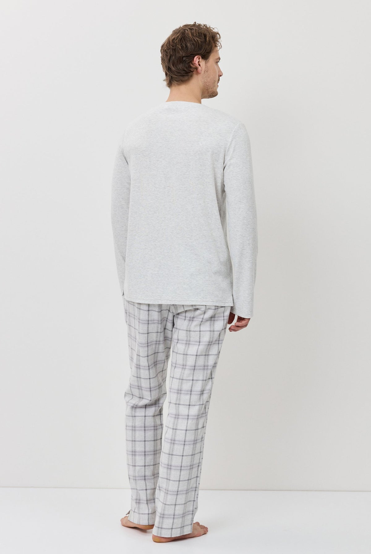 Man wearing a light gray sweater and checkered pants standing against a white background