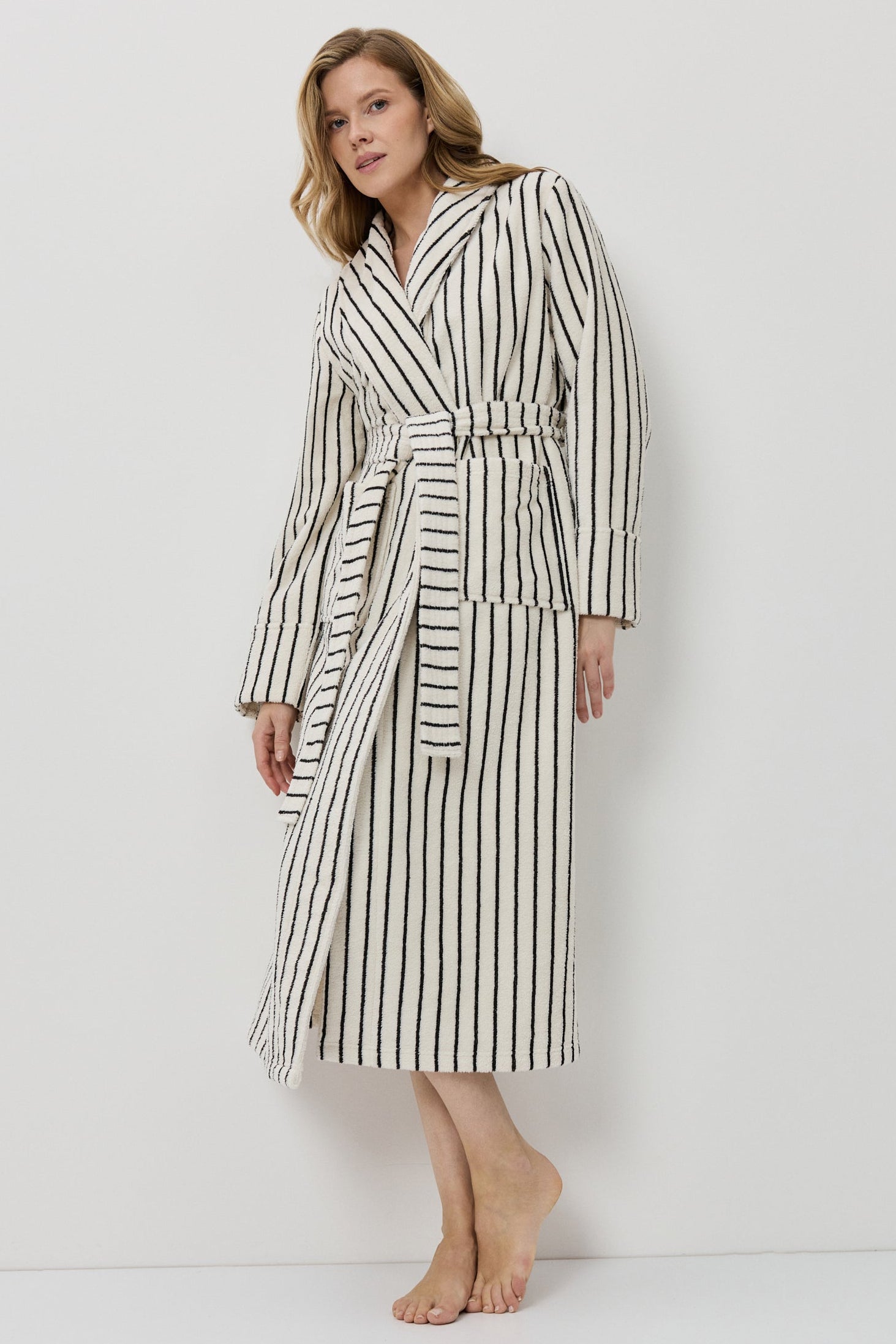 A woman modeling a long-sleeved, knee-length striped bathrobe with a matching tie belt, standing against a plain white studio background.