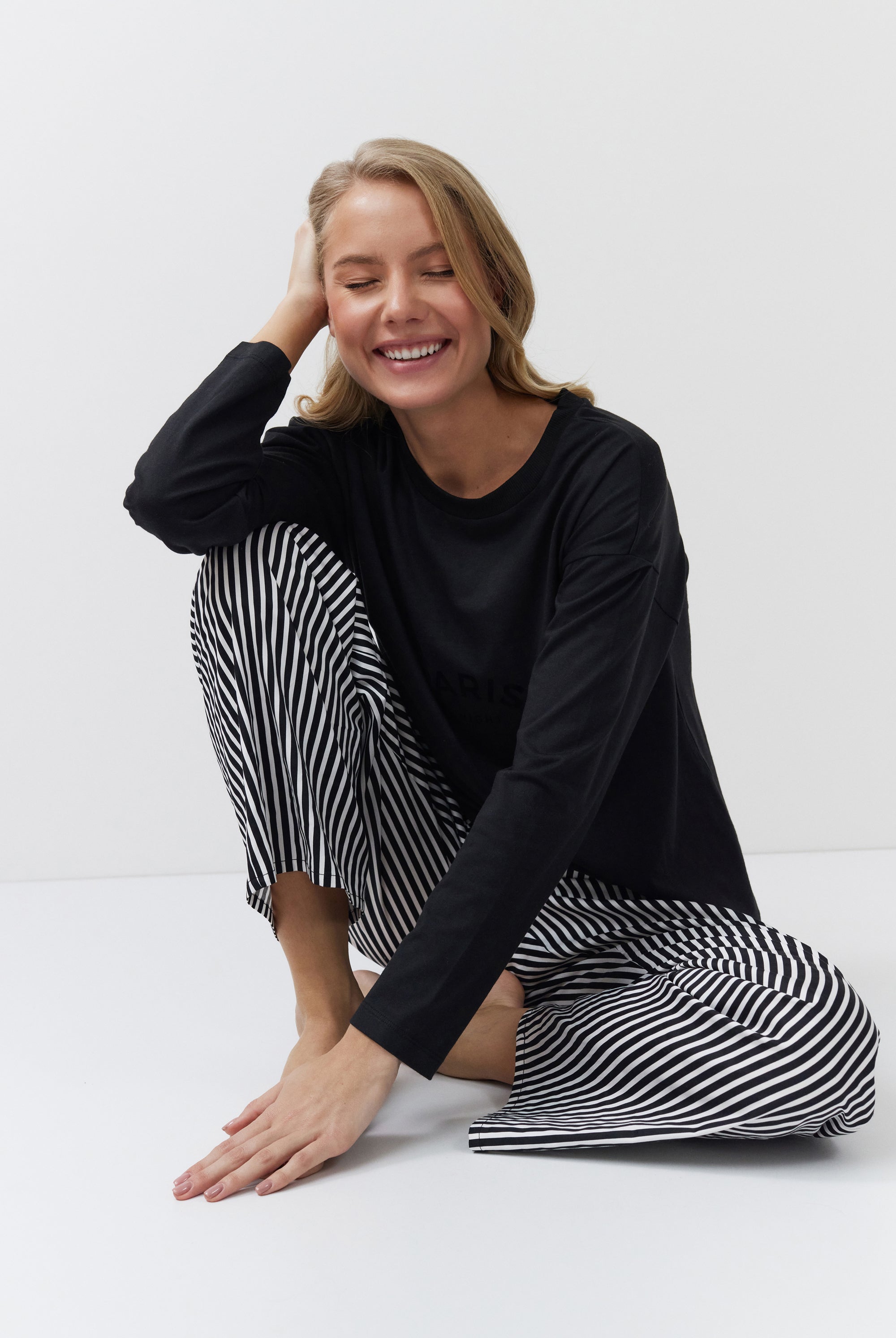 Woman wearing a black long-sleeve top and black and white striped pants sitting on a white floor.