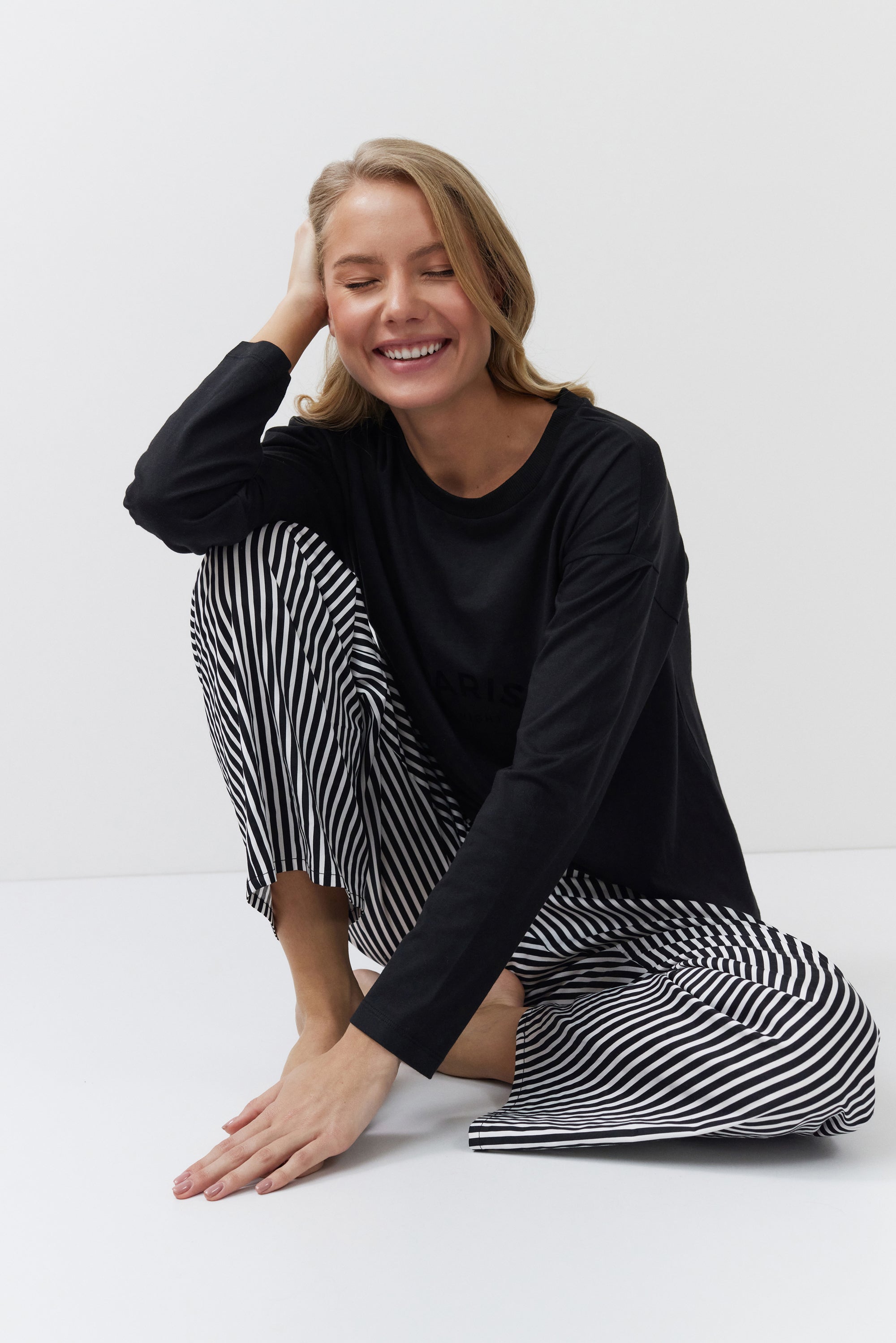 Woman wearing a black long-sleeve top and black and white striped pants sitting on a white floor.