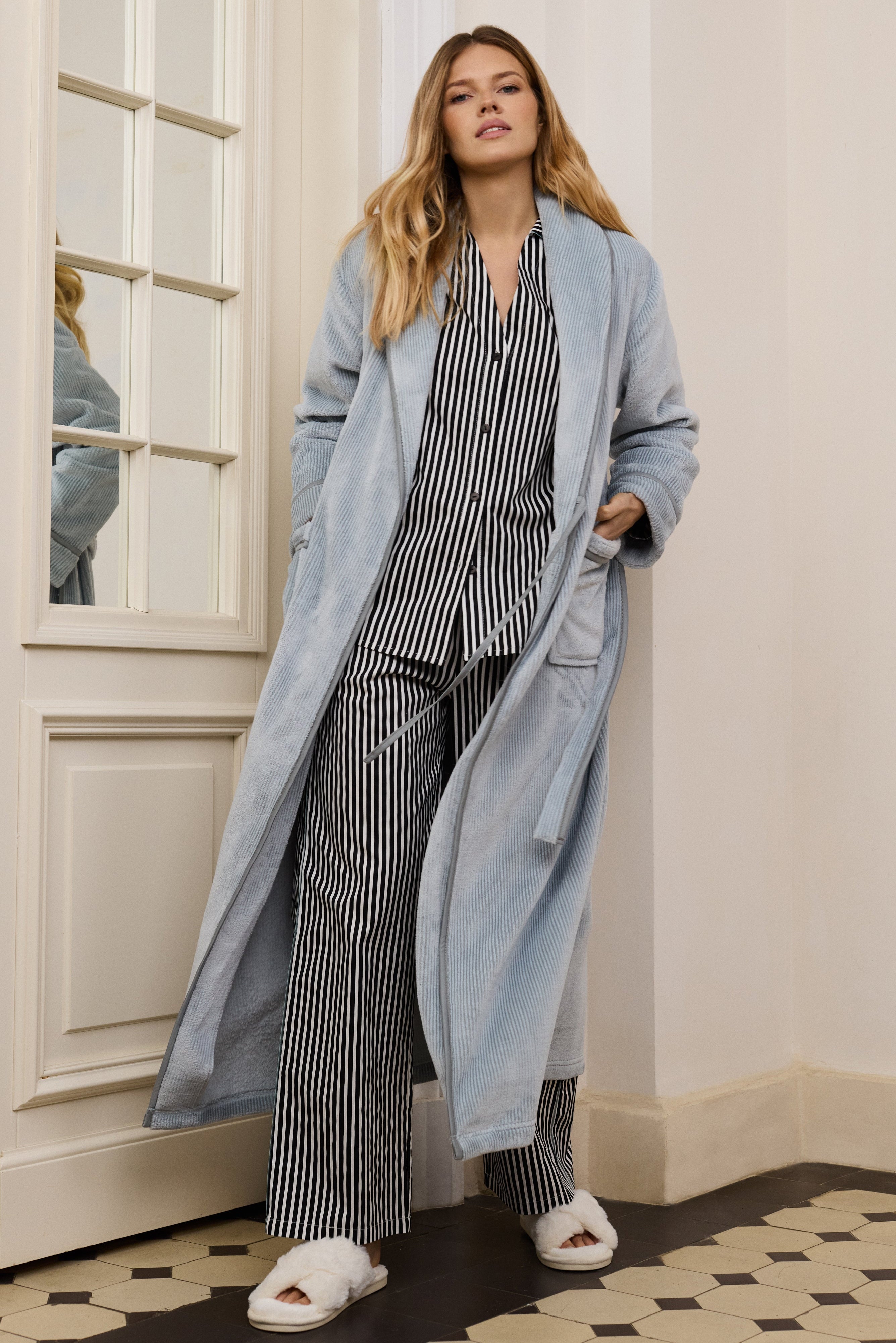 Woman wearing a long light blue bathrobe over a striped outfit in a room with white walls and a patterned floor.
