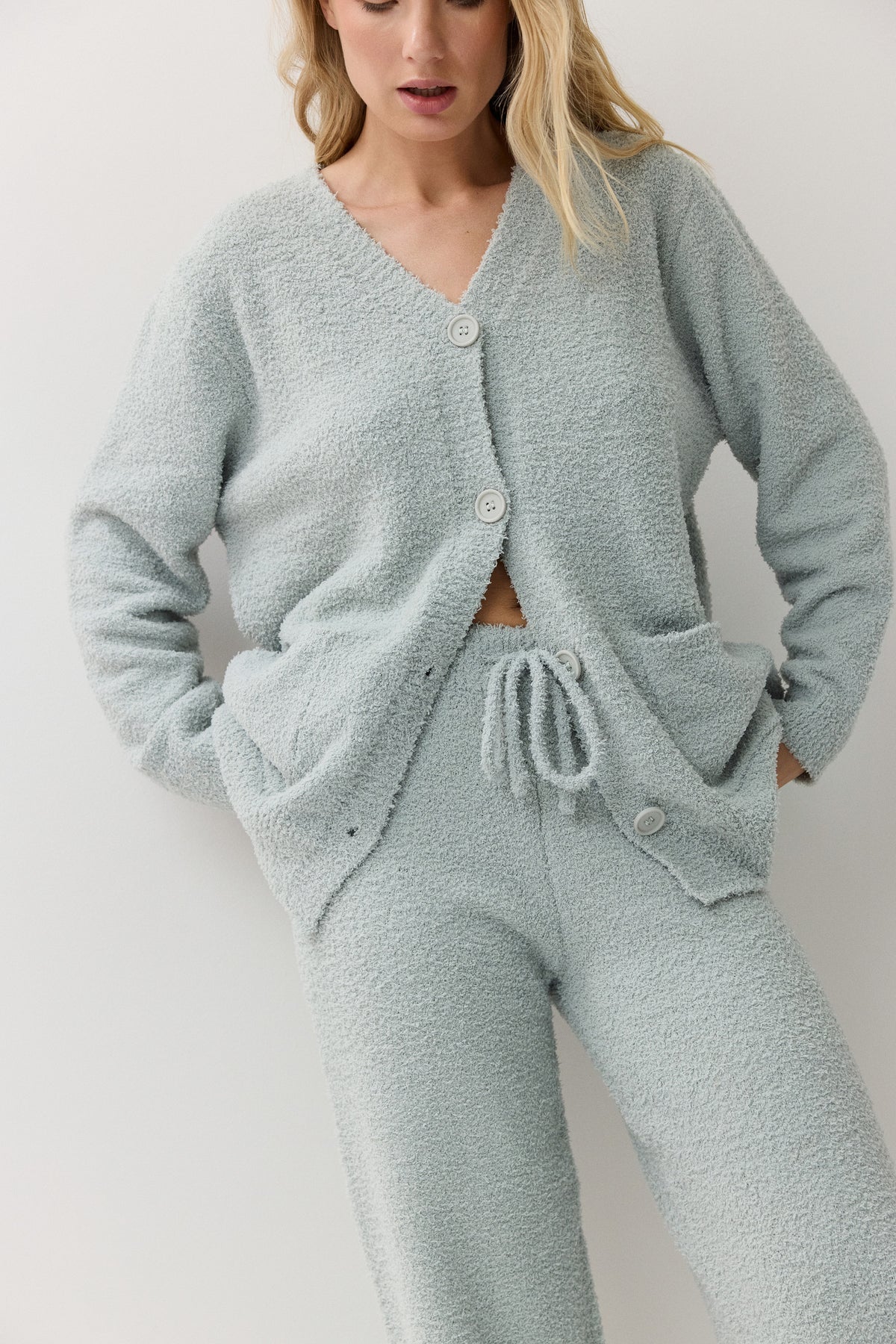 A woman wearing a light sage green fuzzy knit loungewear set, consisting of a cardigan and matching pants, against a plain light gray background.