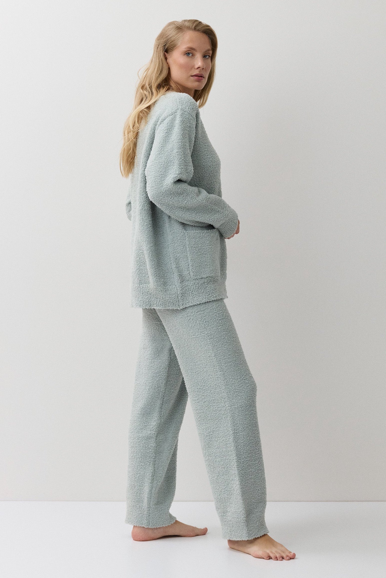 A woman stands barefoot in a studio wearing a matching light sage green loungewear set consisting of a long-sleeve knit top and wide-leg pants.