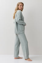 A woman stands barefoot in a studio wearing a matching light sage green loungewear set consisting of a long-sleeve knit top and wide-leg pants.