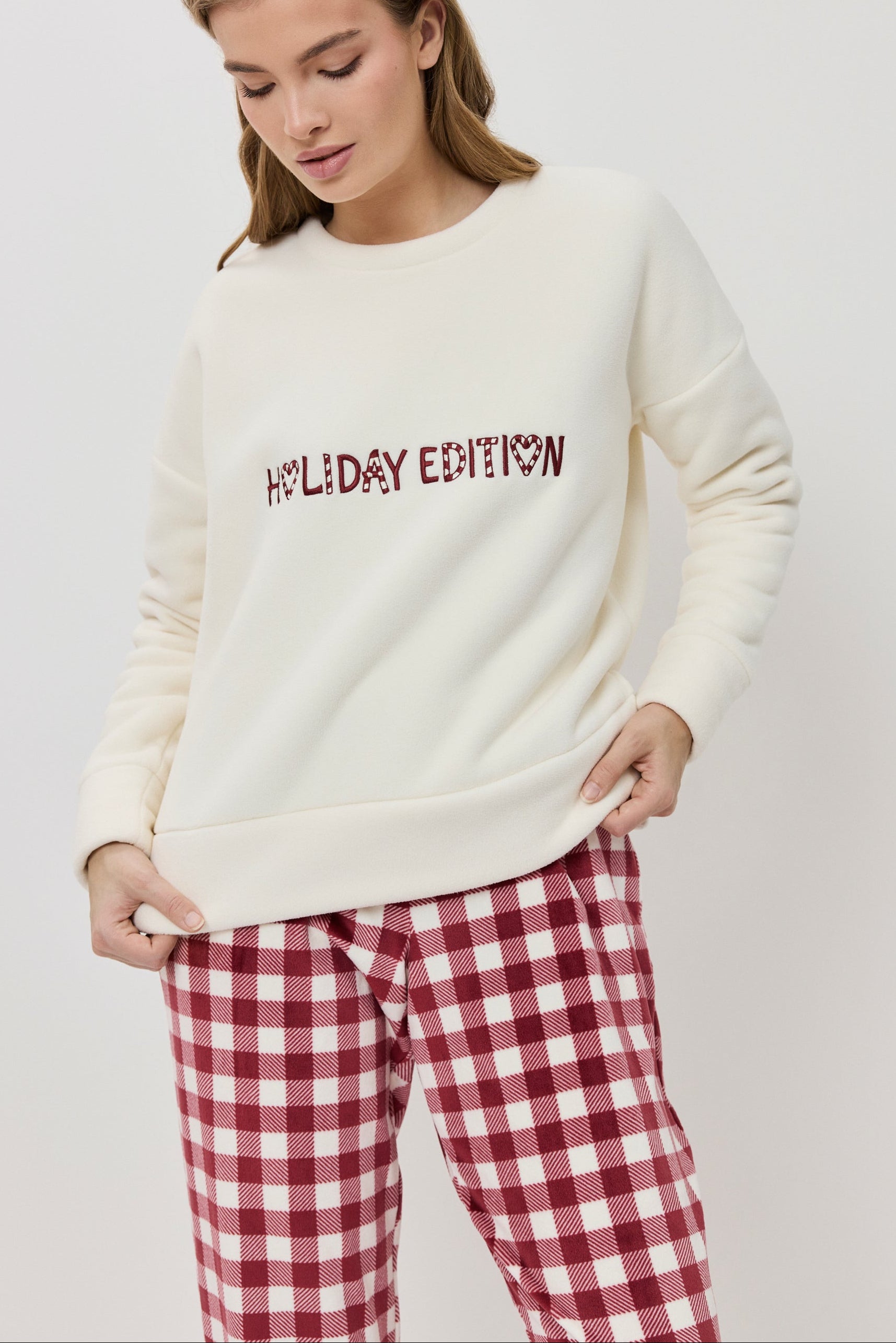 Woman wearing a cream sweater with 'Holiday Edition' text and red plaid pants on a white background