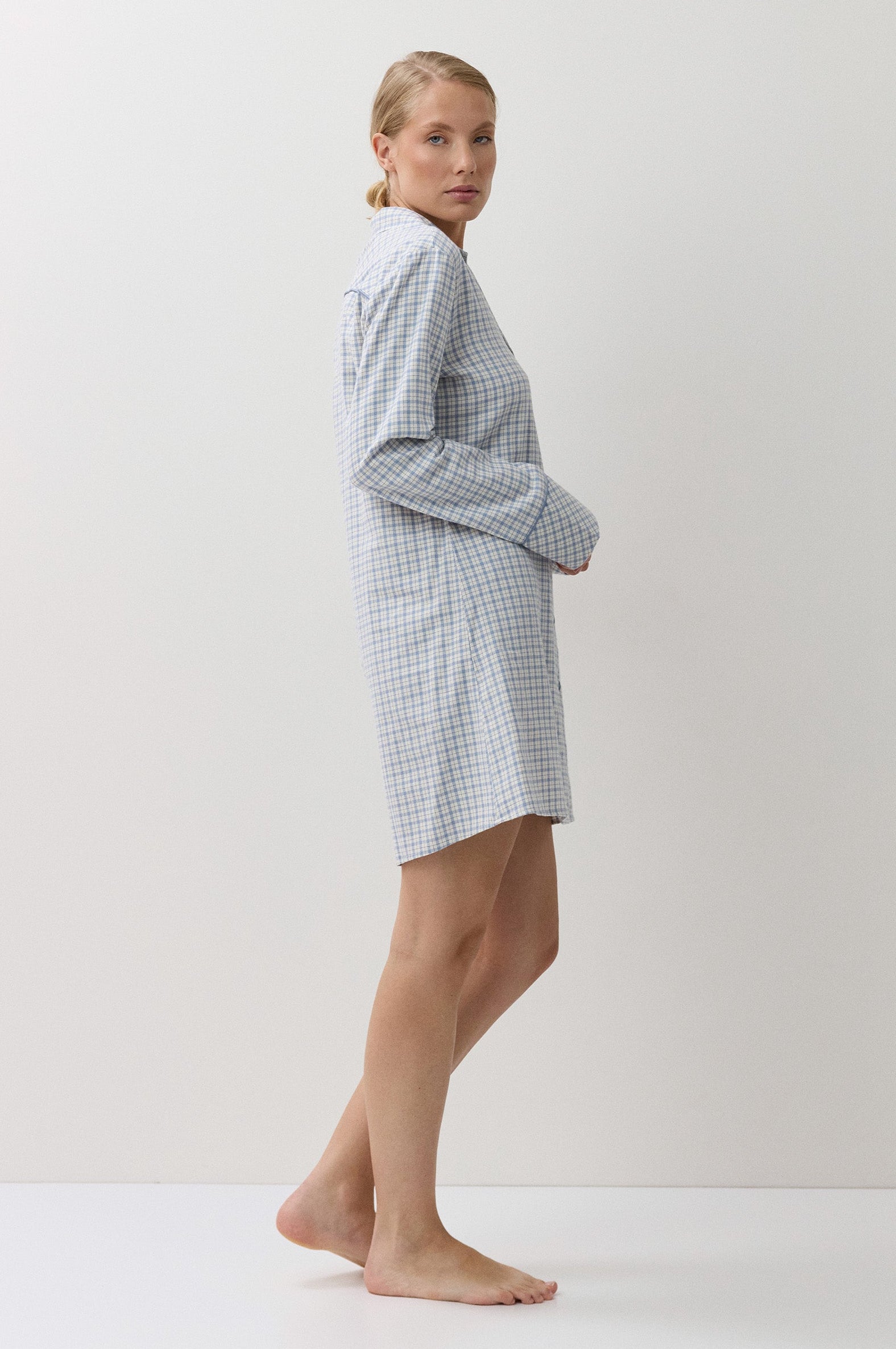 A woman stands barefoot in profile wearing a light blue and white gingham check long-sleeve robe against a plain off-white studio background.