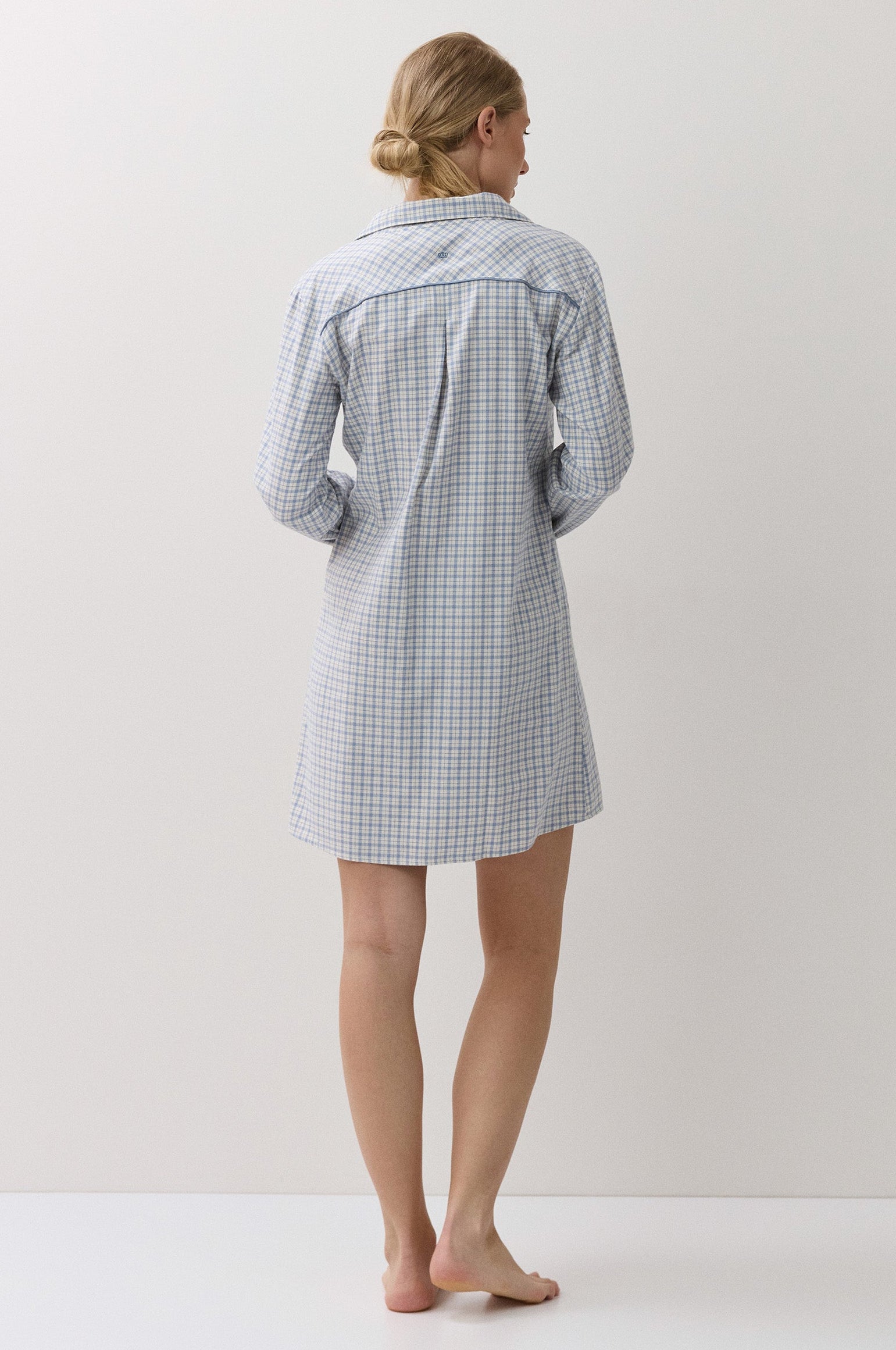 A woman stands barefoot from the back wearing a light blue and white gingham check long-sleeve shirt dress or pajama shirt against a plain off-white studio background.