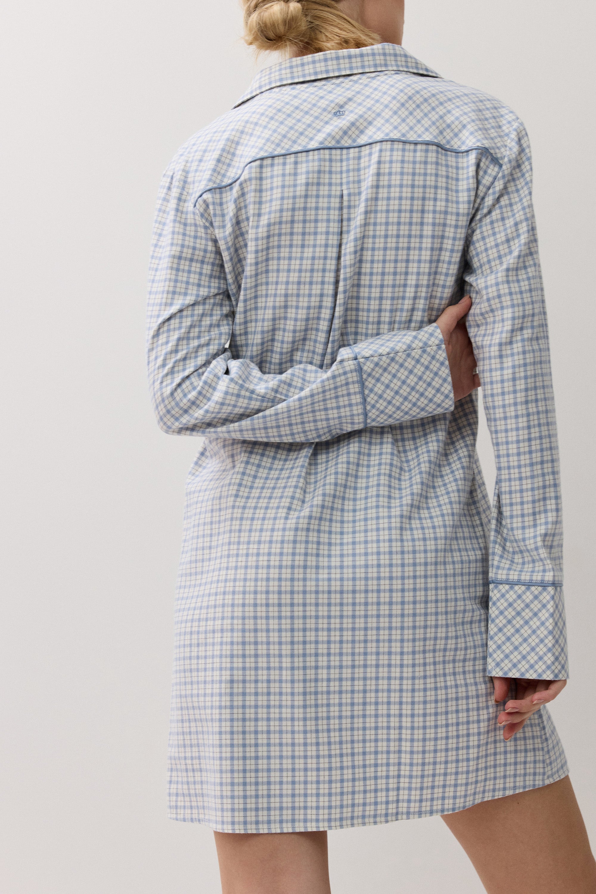 A woman shown from the back wearing a light blue and white gingham plaid shirt dress with matching piping details on the collar, cuffs, and back seams, set against a plain light gray studio background.