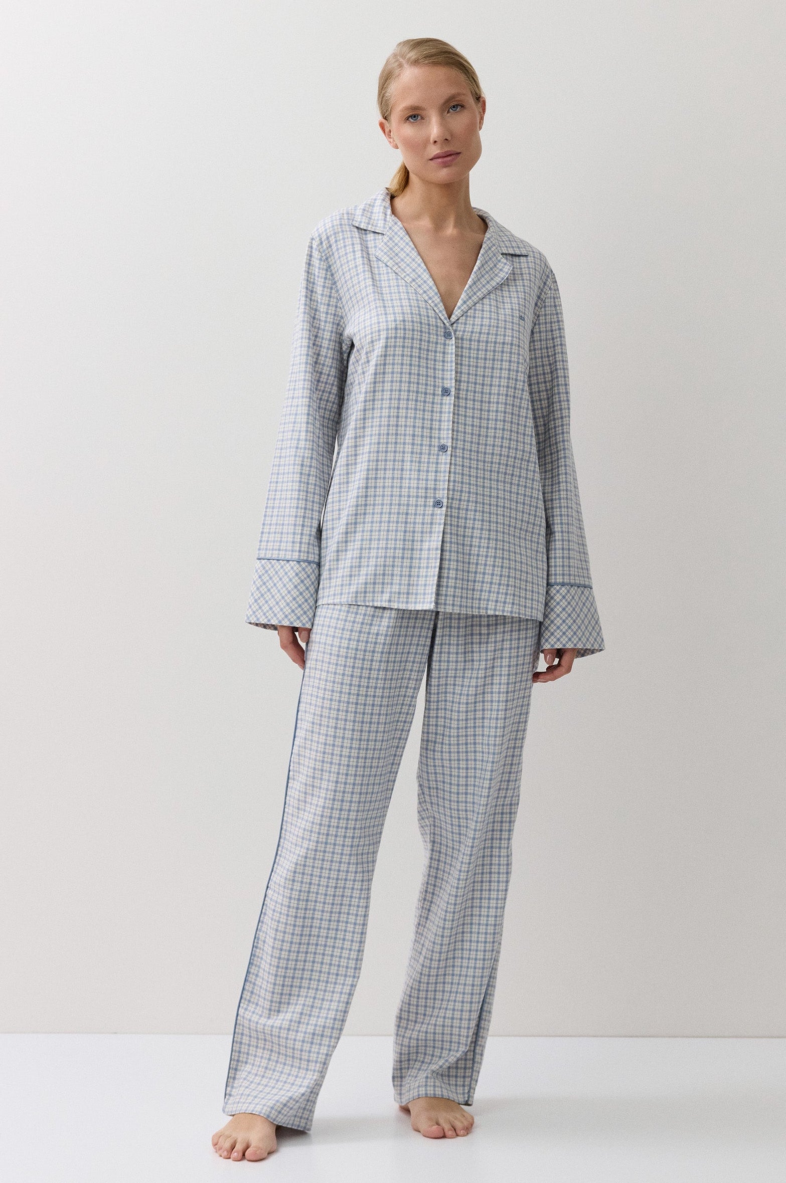 A woman stands barefoot in a studio wearing a matching long-sleeve button-front pajama top and wide-leg pajama pants in a small blue and white gingham check pattern.
