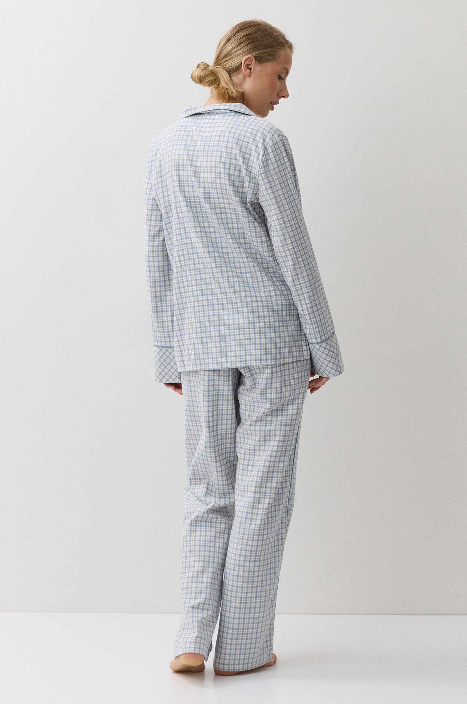 A woman stands with her back to the camera wearing a light blue and white gingham pajama set, consisting of a long-sleeve button-up top and matching pants, in a minimalist studio setting.
