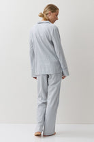 A woman stands with her back to the camera wearing a light blue and white gingham pajama set, consisting of a long-sleeve button-up top and matching pants, in a minimalist studio setting.