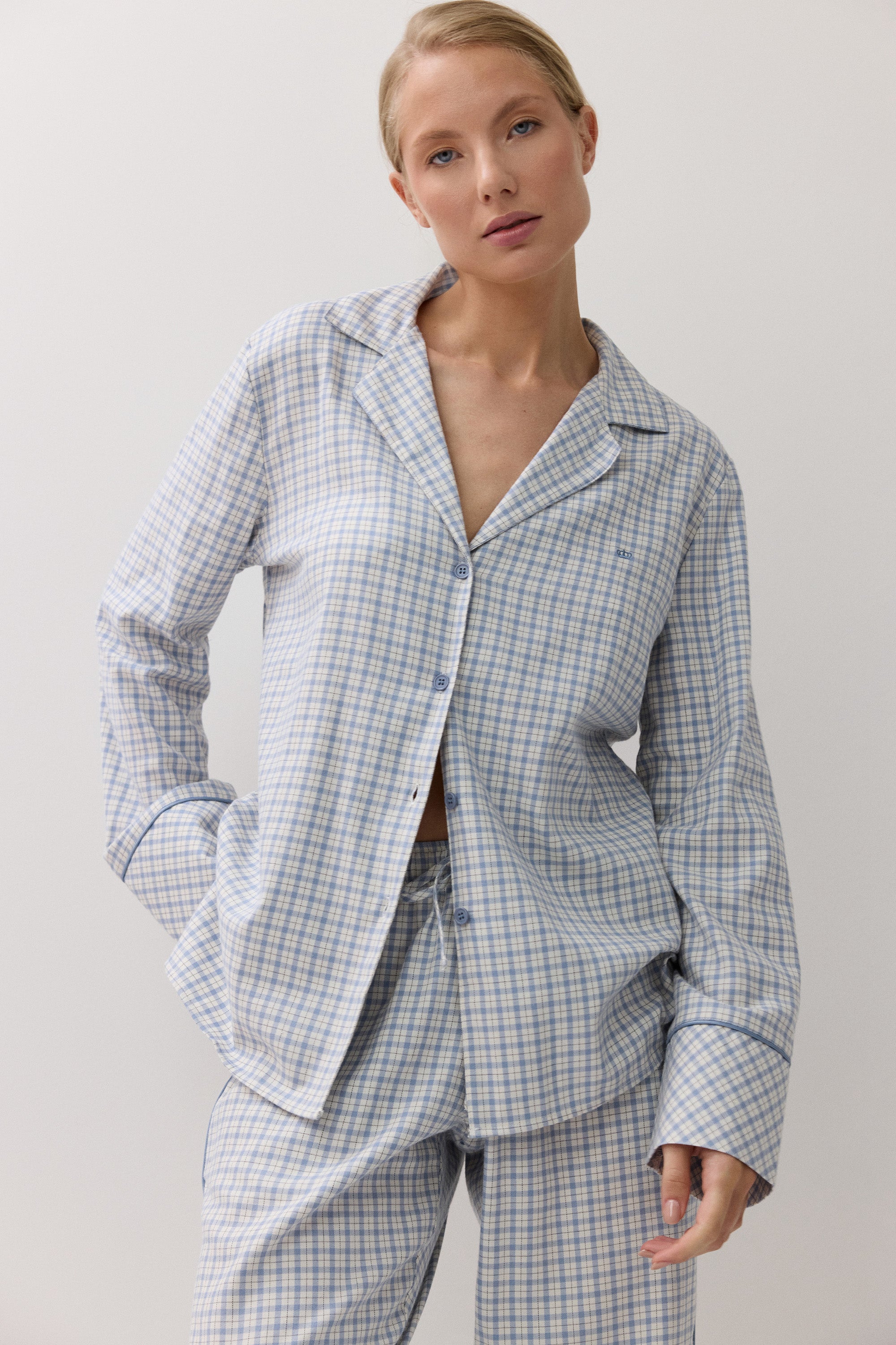 A woman modeling a light blue and white checkered pajama set, featuring a long-sleeve button-up shirt and matching pants, against a plain white background.