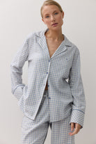 A woman modeling a light blue and white checkered pajama set, featuring a long-sleeve button-up shirt and matching pants, against a plain white background.