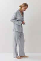 A woman wearing a light blue and white gingham pajama set, standing barefoot against a plain white studio background.