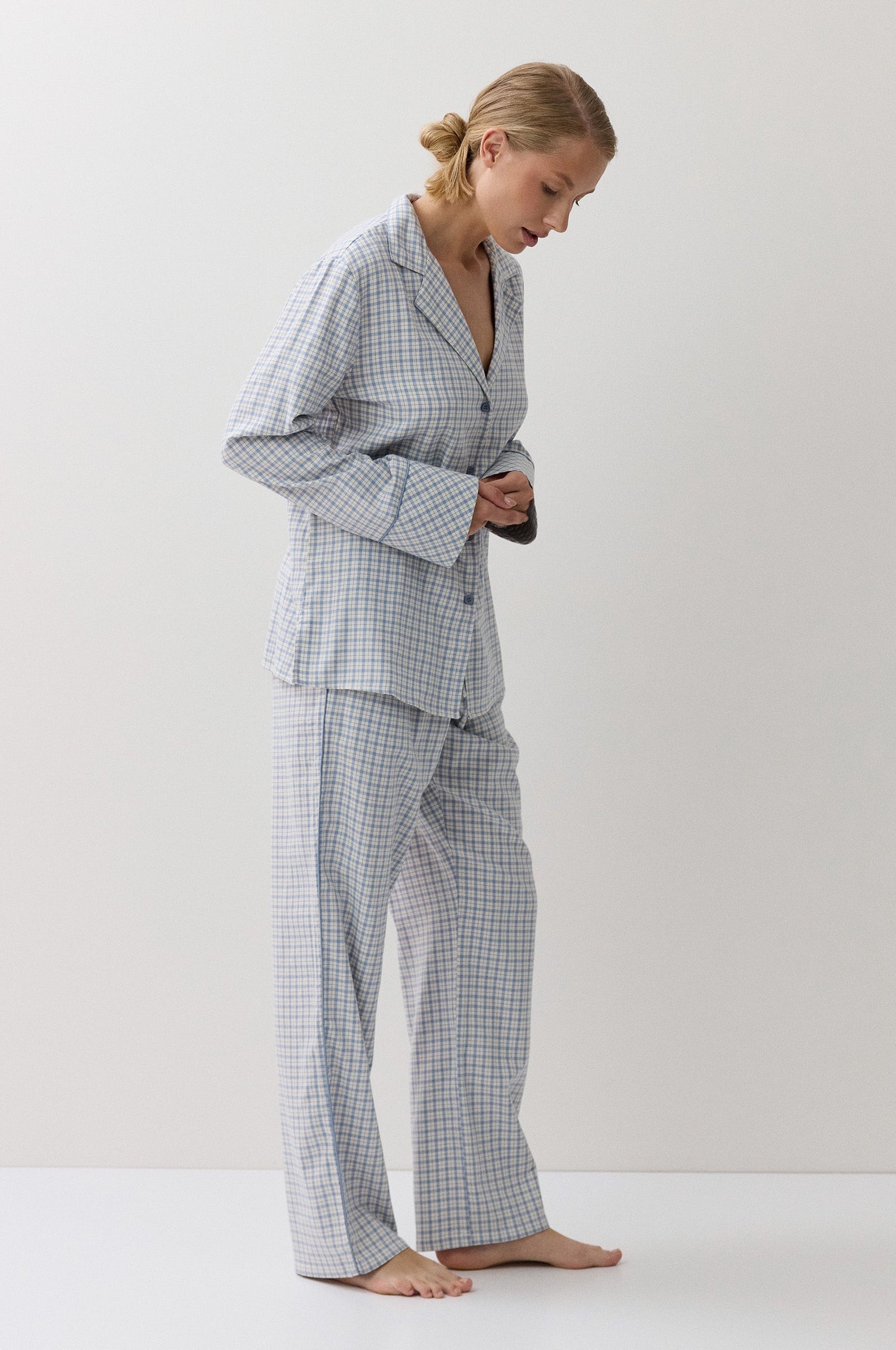 A woman wearing a light blue and white gingham pajama set, standing barefoot against a plain white studio background.