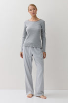 A woman stands barefoot in a studio wearing a light gray long-sleeve top and matching gray-and-white gingham pajama pants against a plain light background.