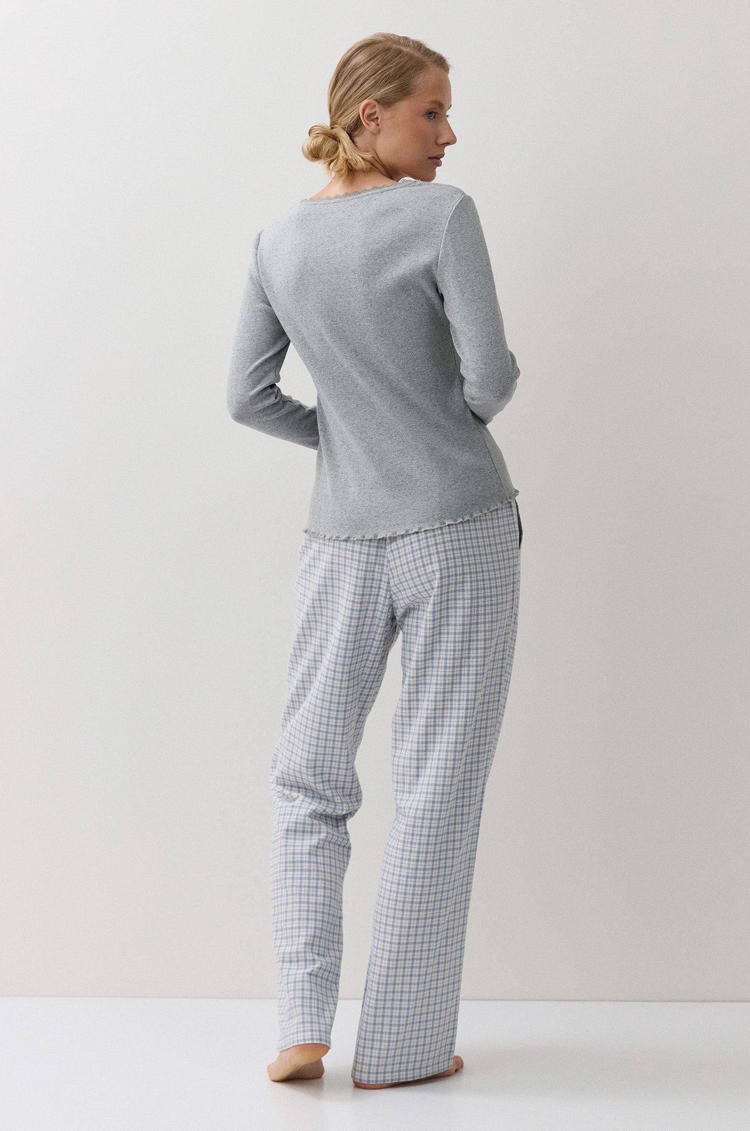A woman stands barefoot against a plain white studio background, viewed from the back, wearing a light gray long-sleeve top and matching white-and-blue checkered pajama pants.